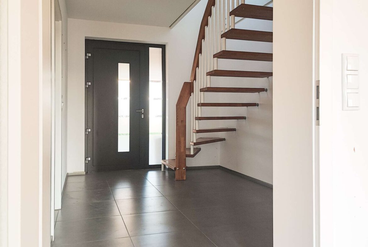 Modern entry hall with a dark front door featuring vertical glass panels, grey tile floor, and a wooden staircase with white railing on the right.