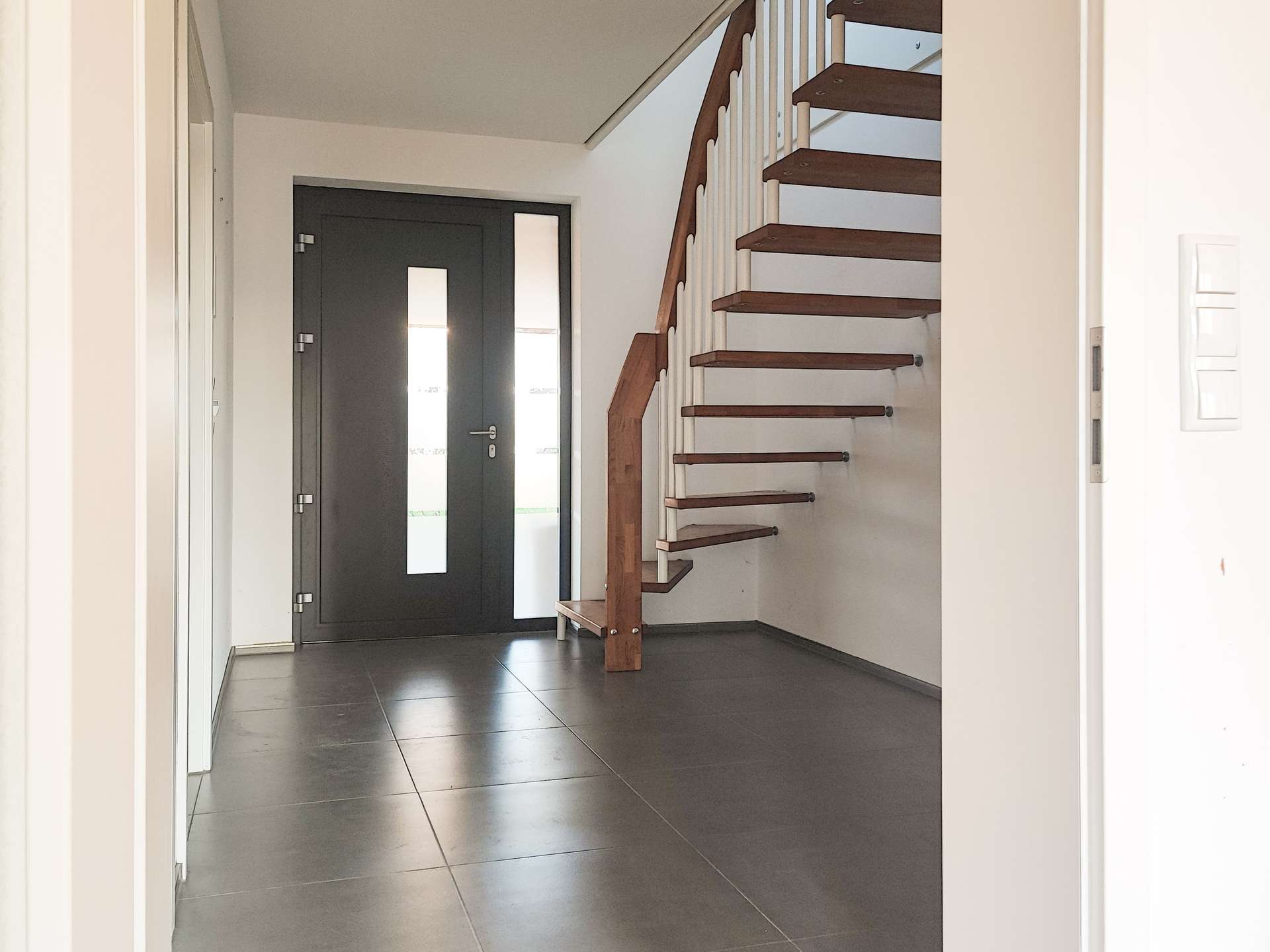 Modern entry hall with a dark front door featuring vertical glass panels, grey tile floor, and a wooden staircase with white railing on the right.