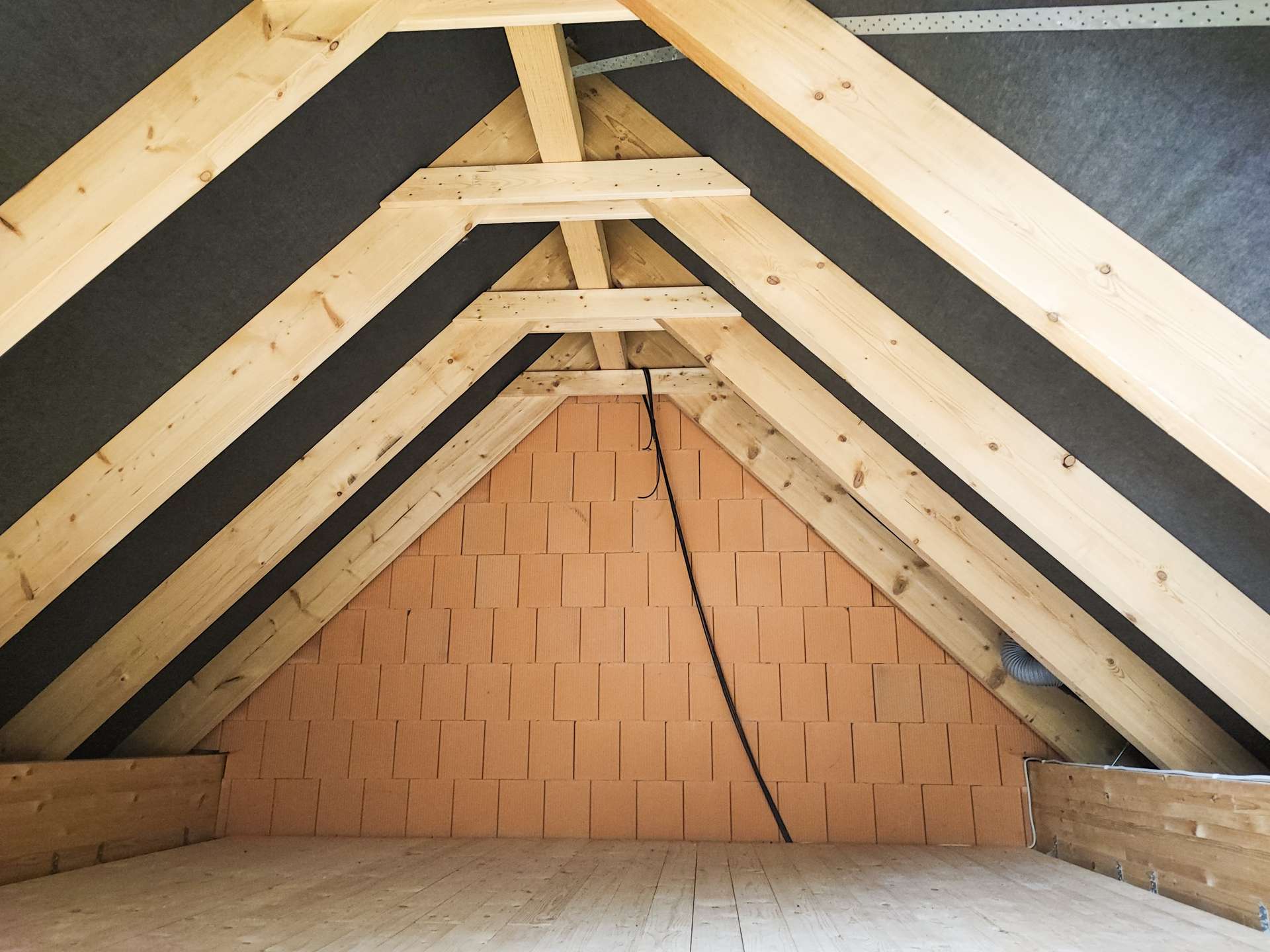 Attic with exposed wooden roof trusses, orange brick back wall, and a black cable running down the center.