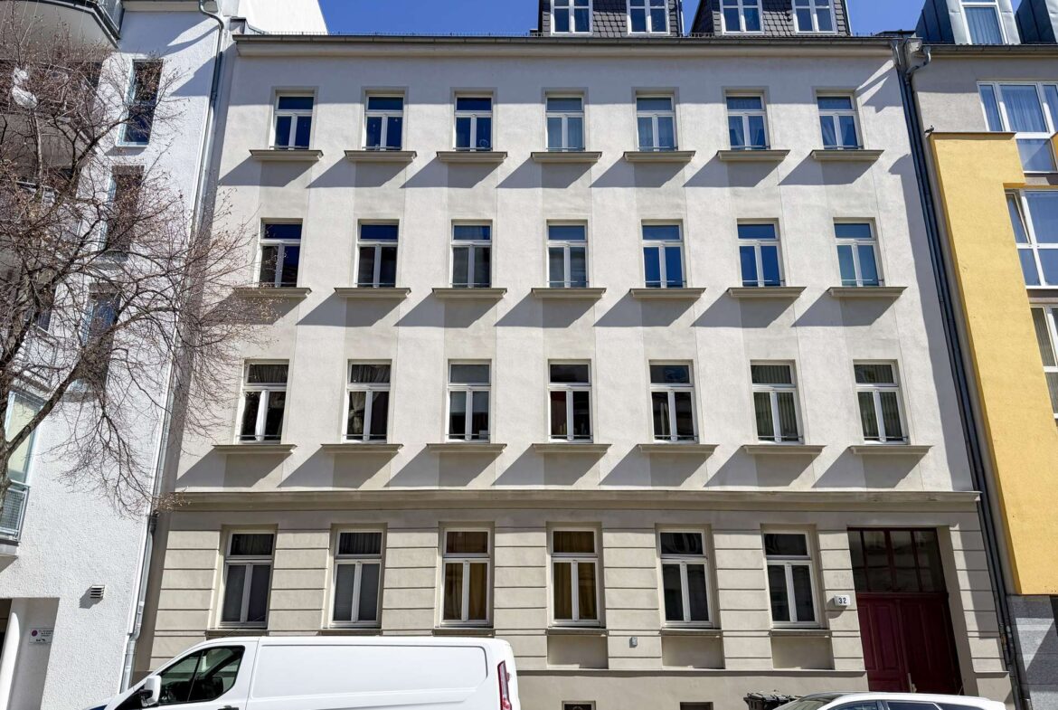 Front view of a pale beige apartment building with evenly spaced windows, a white van parked at the curb, and a clear blue sky above.