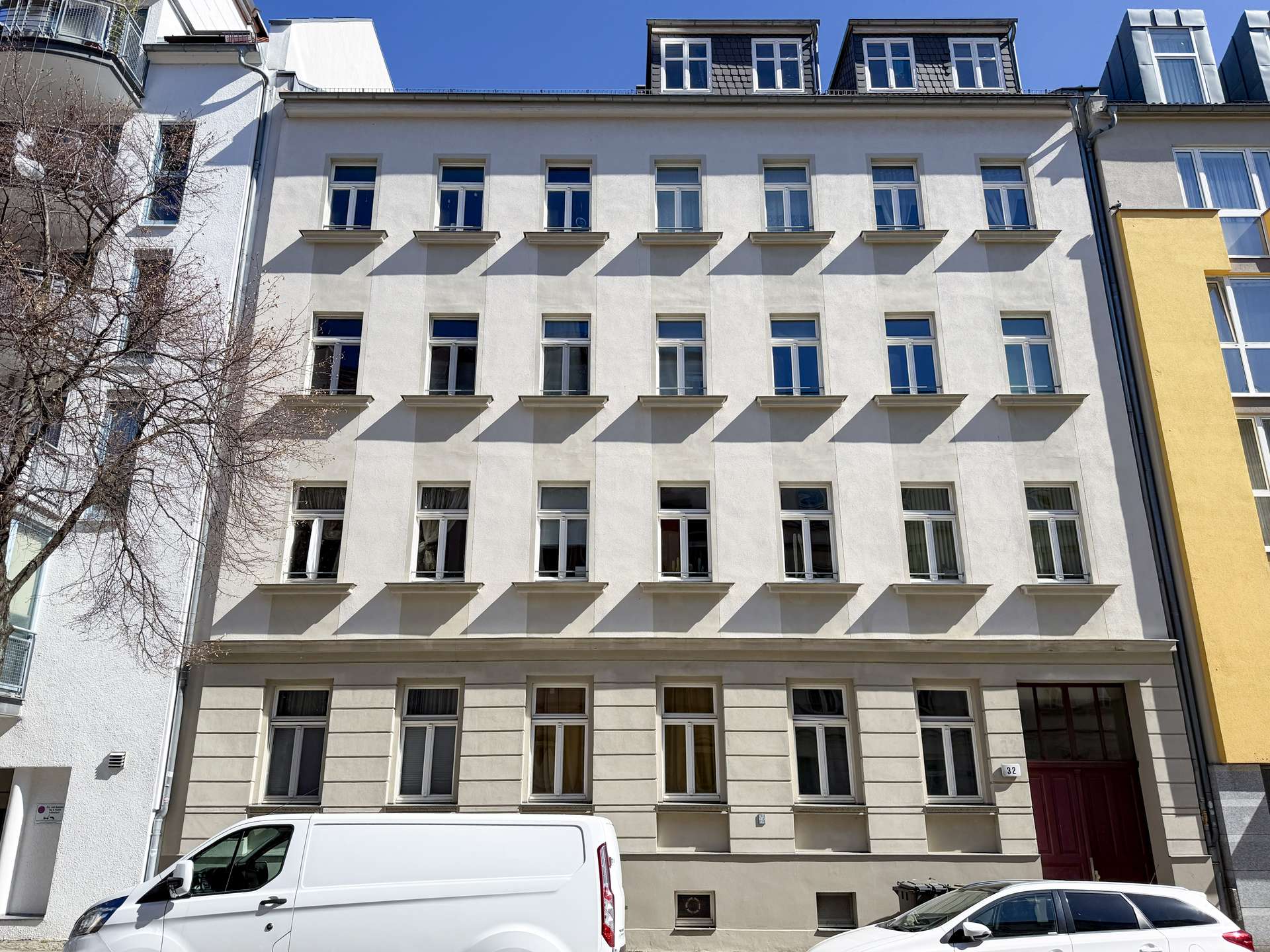 Front view of a pale beige apartment building with evenly spaced windows, a white van parked at the curb, and a clear blue sky above.