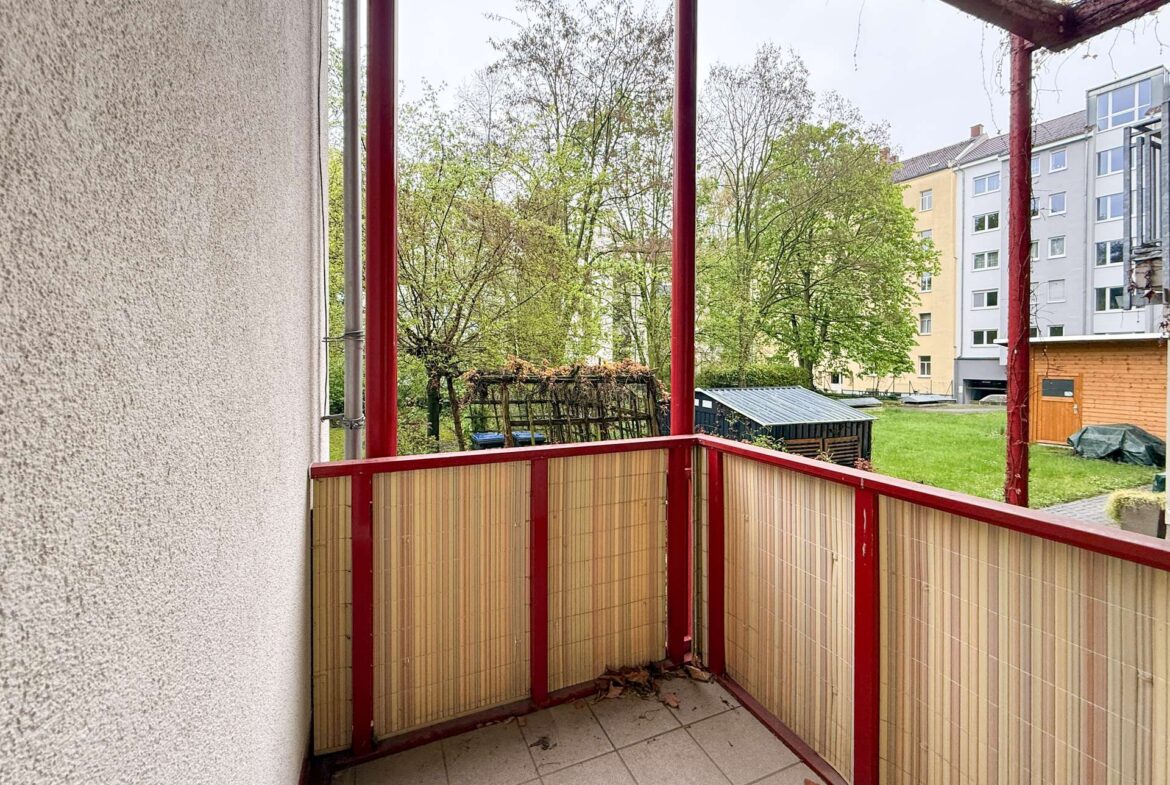 Balcony with red metal rails and woven bamboo privacy screens, looking out at trees and apartment buildings in a courtyard.