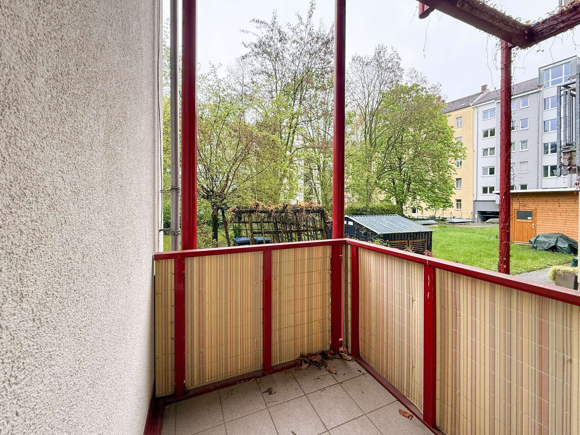 Balcony with red metal rails and woven bamboo privacy screens, looking out at trees and apartment buildings in a courtyard.