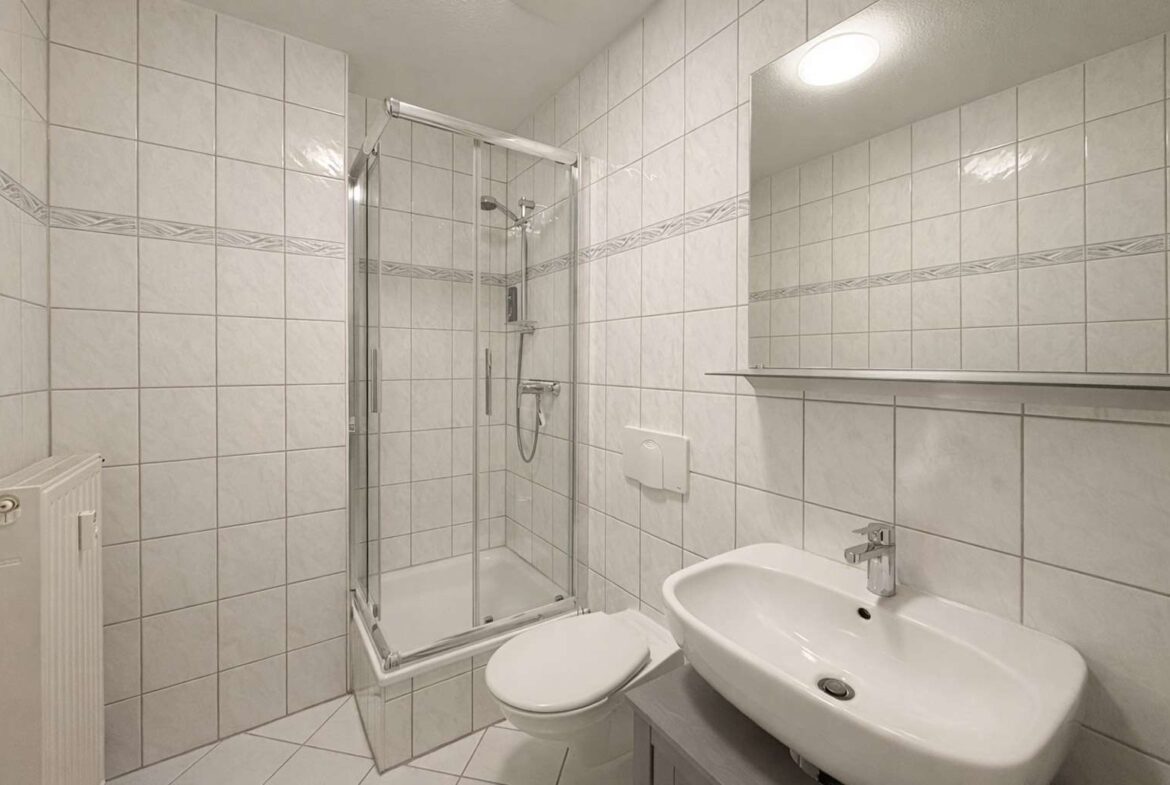 Compact white-tiled bathroom with a glass corner shower, wall-mounted toilet, and oval sink with a shelf above a large mirror.