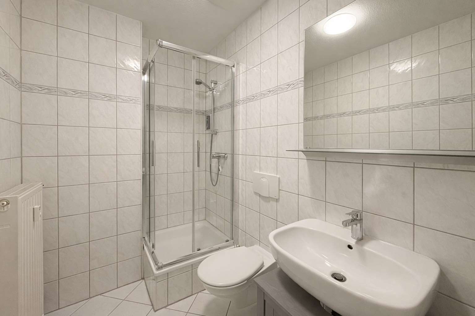Compact white-tiled bathroom with a glass corner shower, wall-mounted toilet, and oval sink with a shelf above a large mirror.