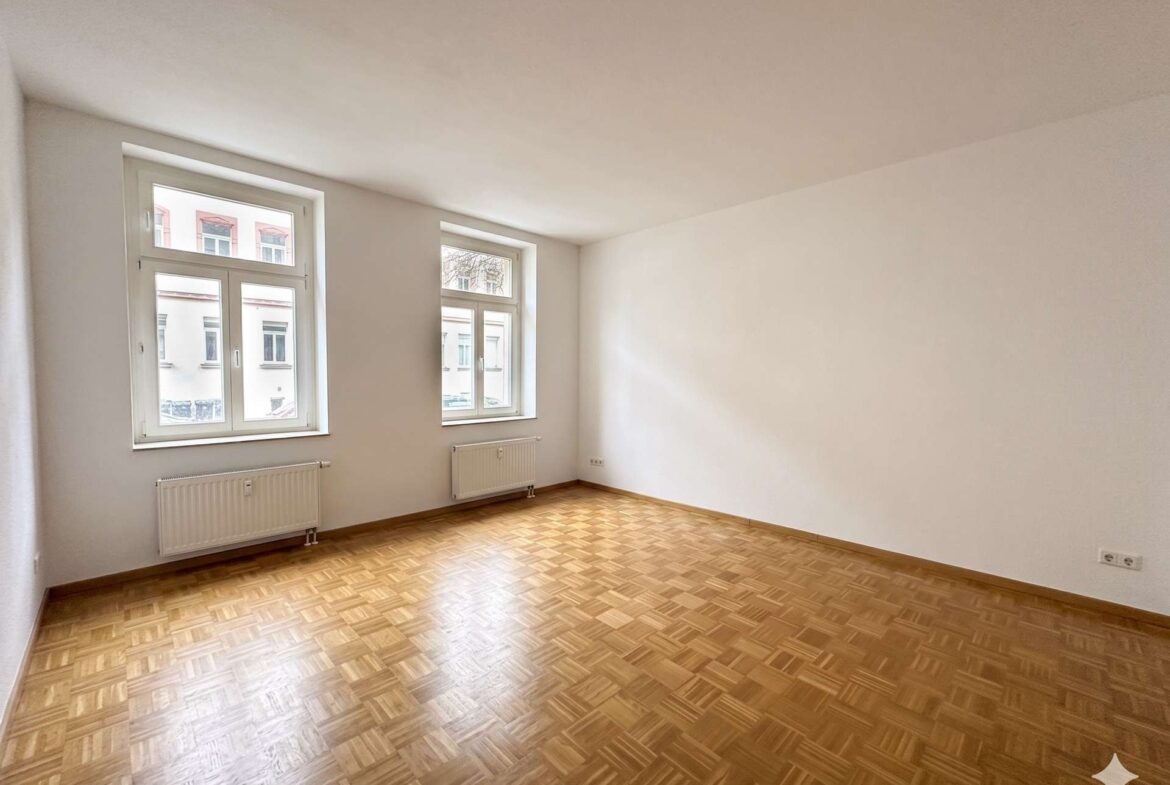 Bright, empty room with two large windows, white walls, and a patterned wood floor (parquet). Radiators along the window wall provide heat.