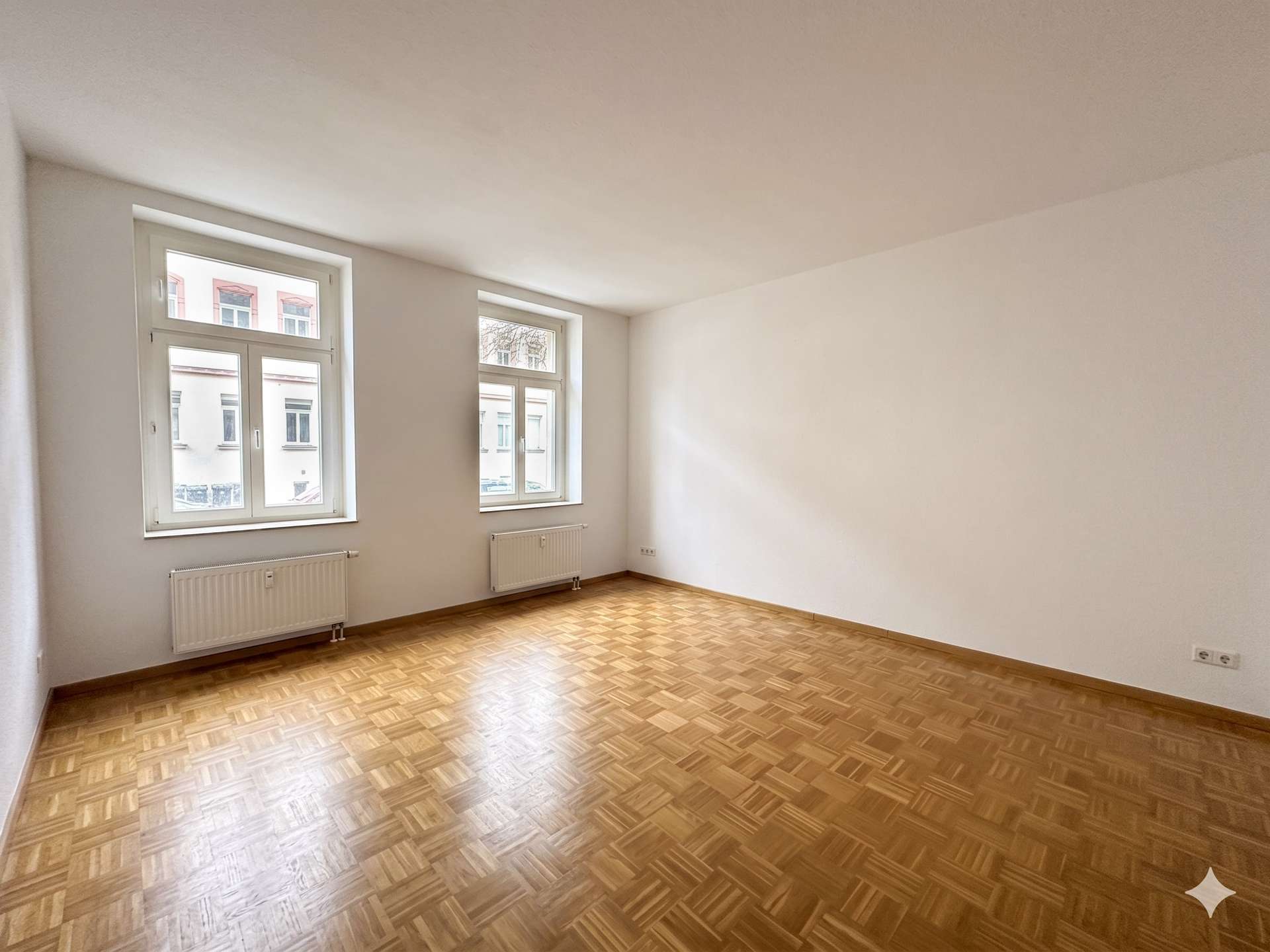 Bright, empty room with two large windows, white walls, and a patterned wood floor (parquet). Radiators along the window wall provide heat.