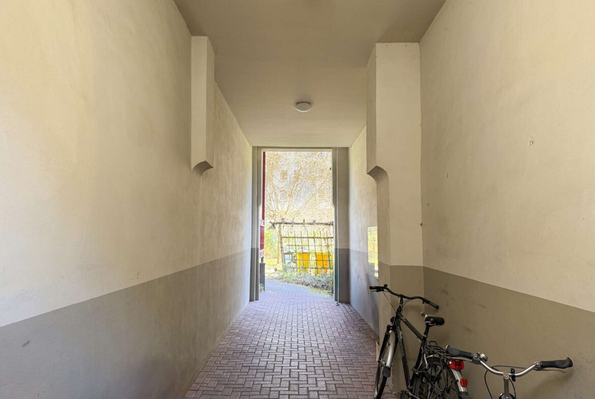 Narrow indoor hallway with bicycles parked along the right wall, leading to an open doorway to a sunny outdoor area.