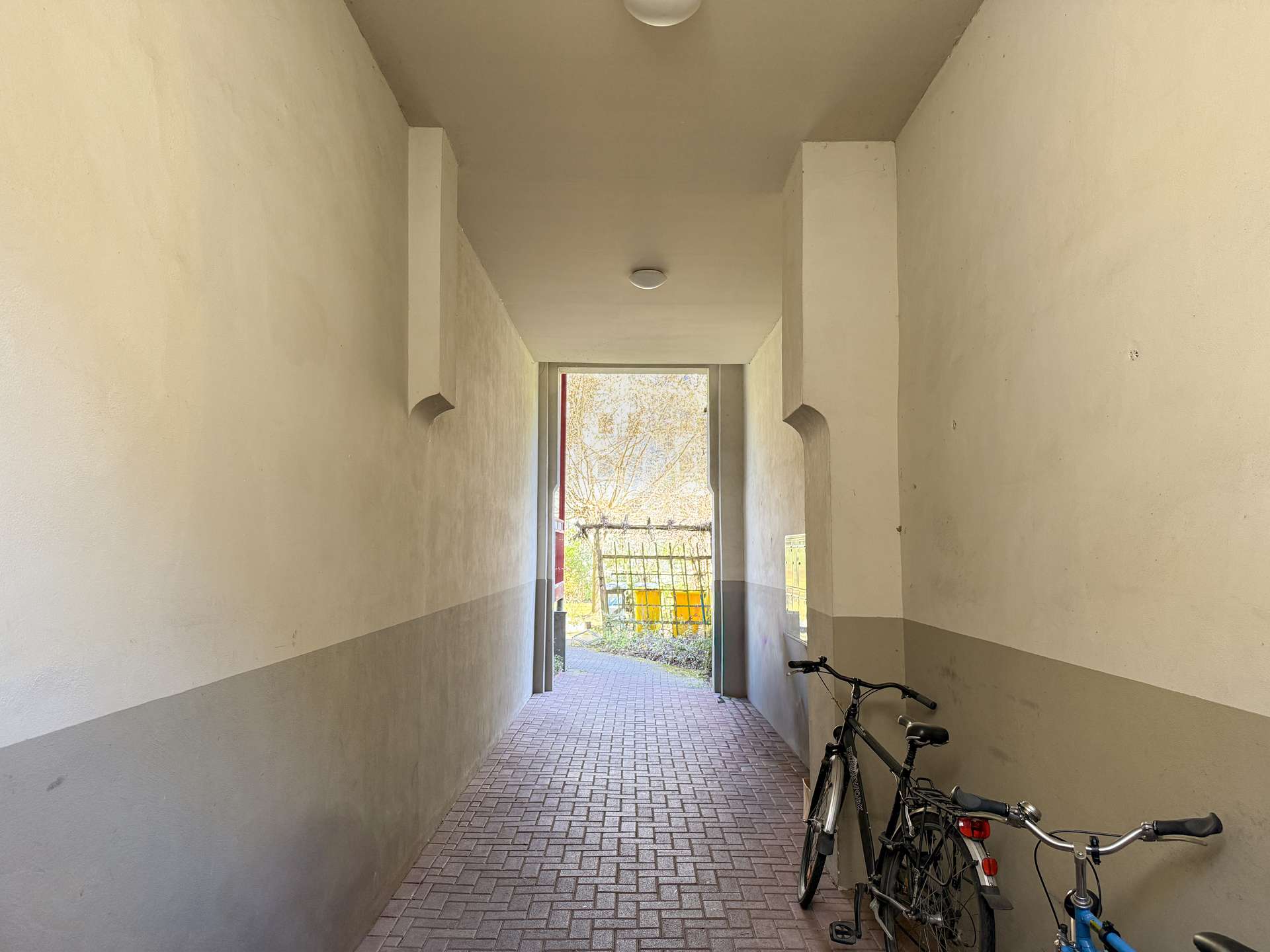 Narrow indoor hallway with bicycles parked along the right wall, leading to an open doorway to a sunny outdoor area.