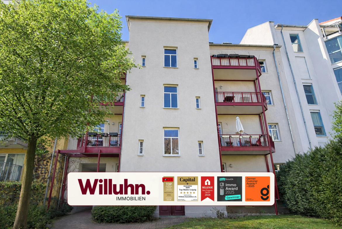 Beige apartment building with red balconies beside a green tree and blue sky; Willuhn Immobilien banner at bottom.