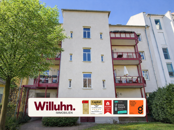 Beige apartment building with red balconies beside a green tree and blue sky; Willuhn Immobilien banner at bottom.