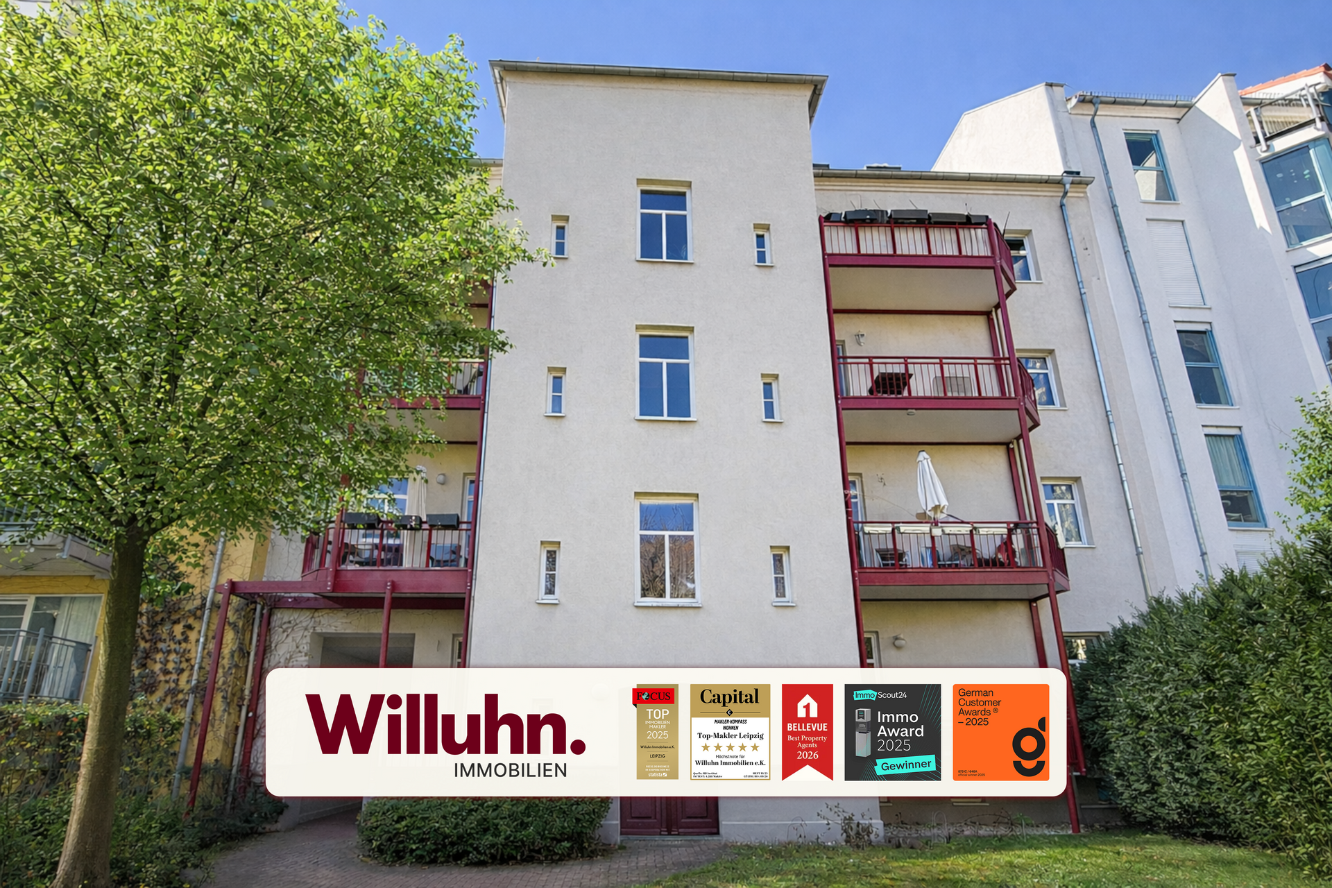 Beige apartment building with red balconies beside a green tree and blue sky; Willuhn Immobilien banner at bottom.