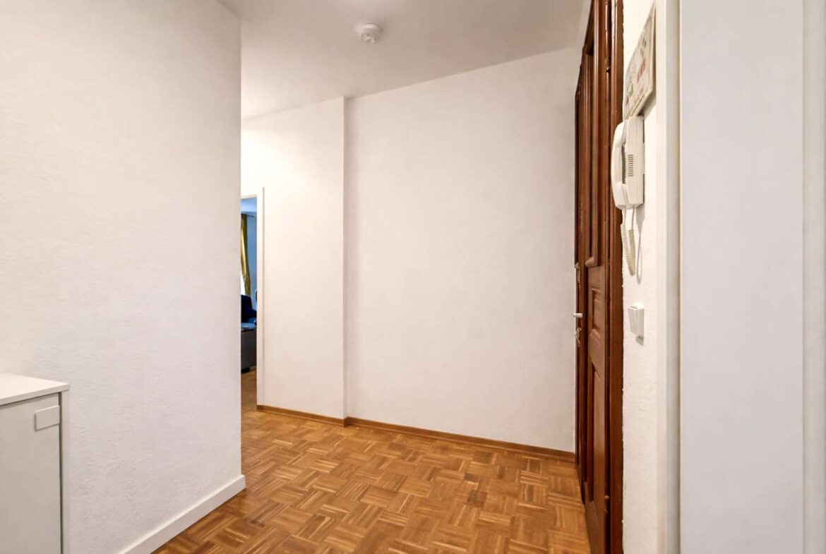 Narrow hallway in a home with parquet floor, white walls, and a wooden door on the right with a wall-mounted intercom beside it.