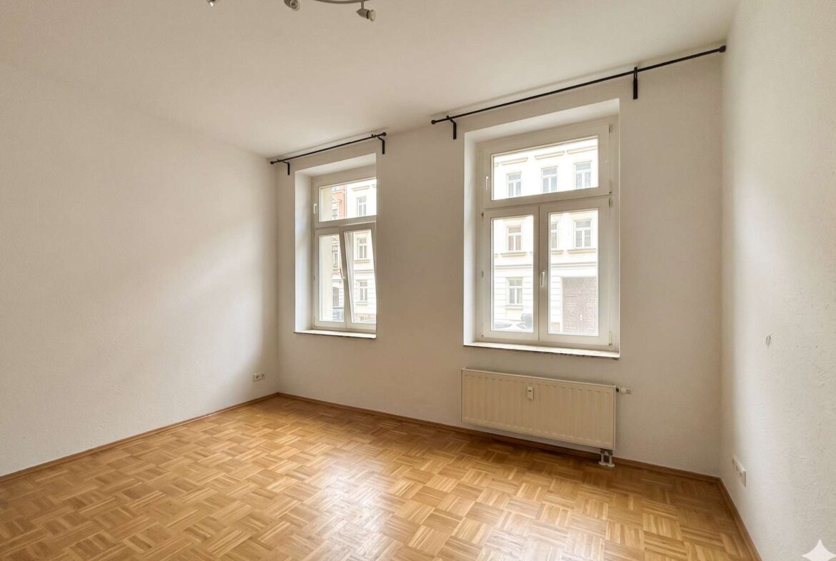 Empty, well-lit room with parquet floor, two large windows, and a wall-mounted radiator beneath the windows.
