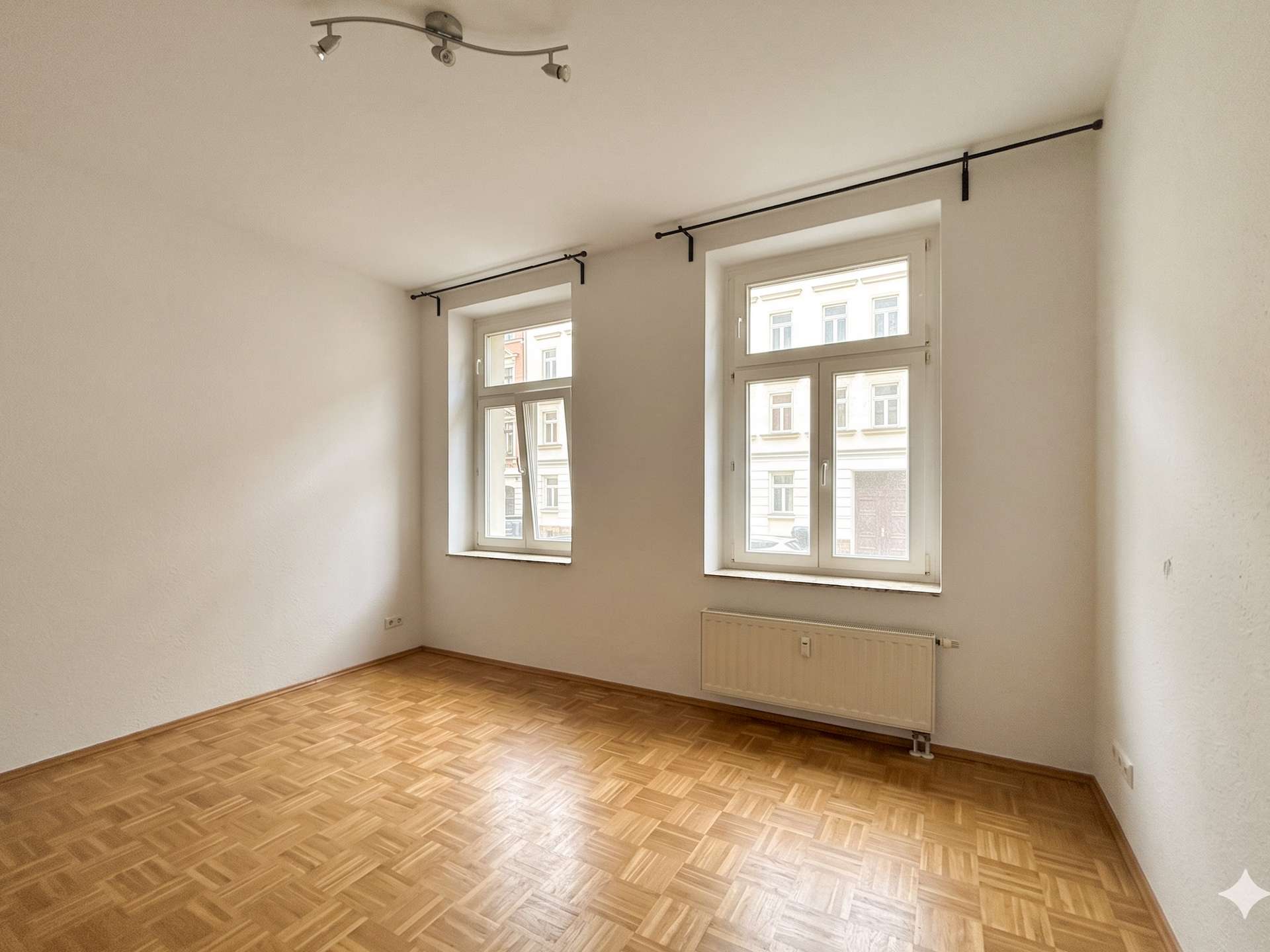 Empty, well-lit room with parquet floor, two large windows, and a wall-mounted radiator beneath the windows.