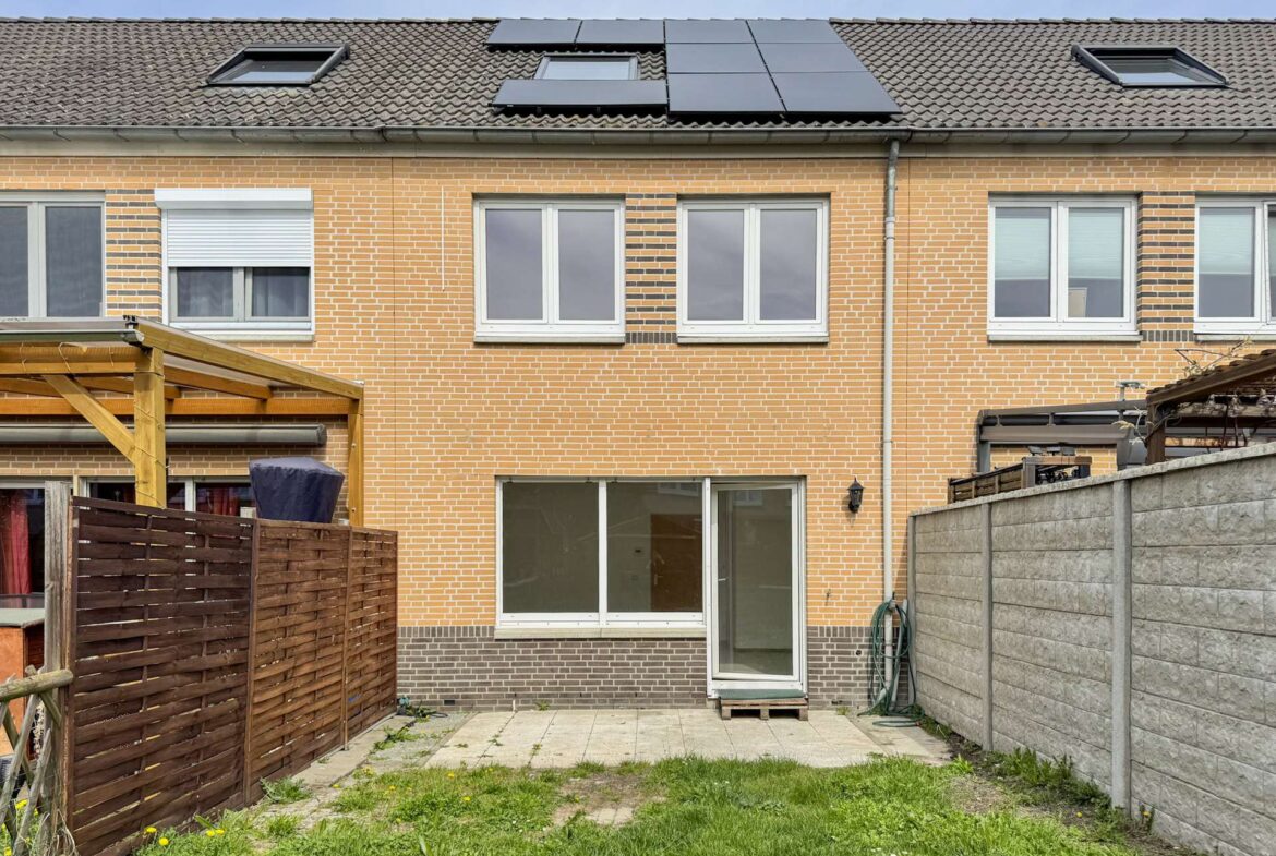 Backyard view of a brick townhouse with a sliding glass door, paved patio, and solar panels on the roof; wooden fence on the left and a concrete privacy wall on the right.