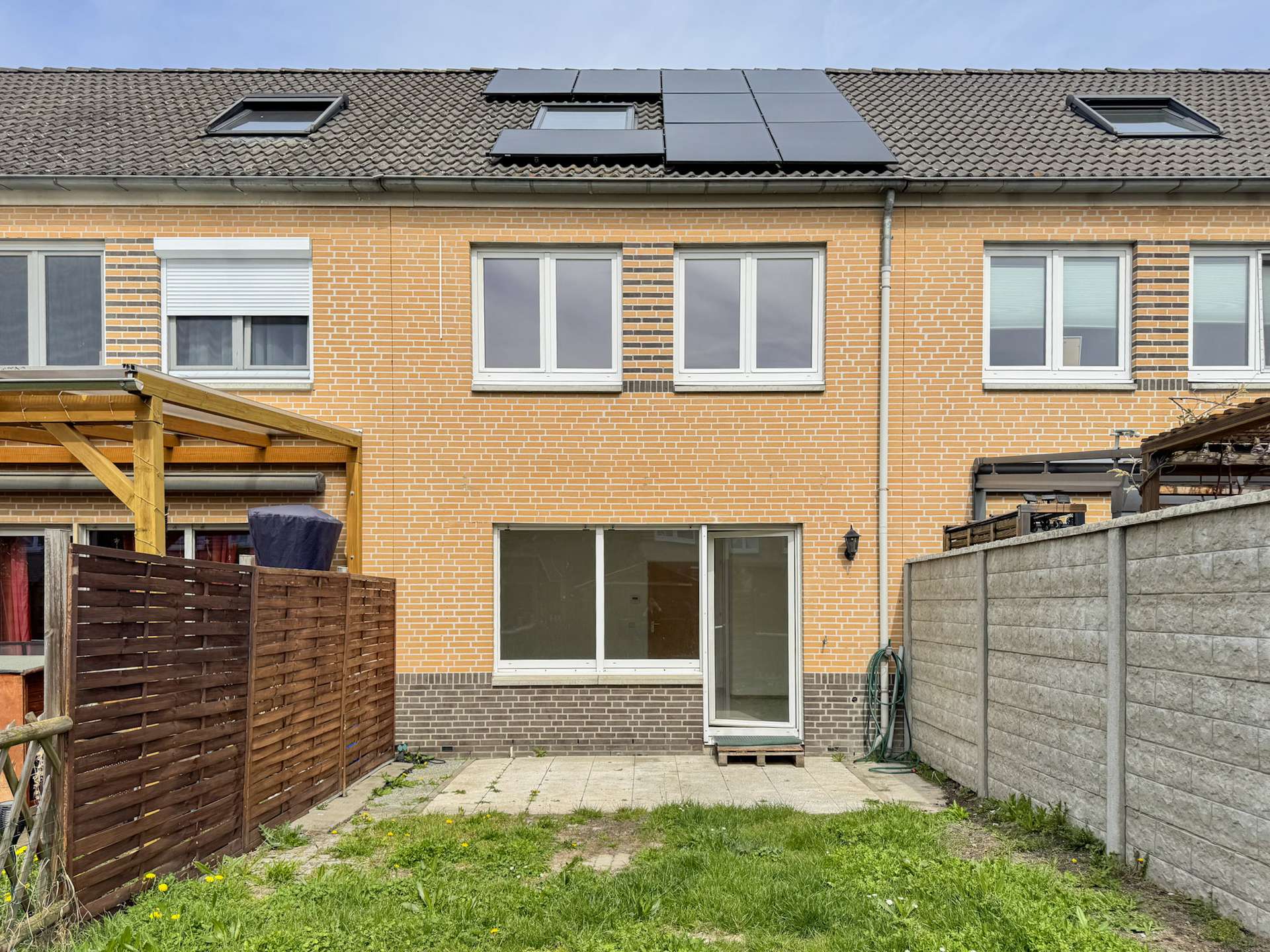 Backyard view of a brick townhouse with a sliding glass door, paved patio, and solar panels on the roof; wooden fence on the left and a concrete privacy wall on the right.