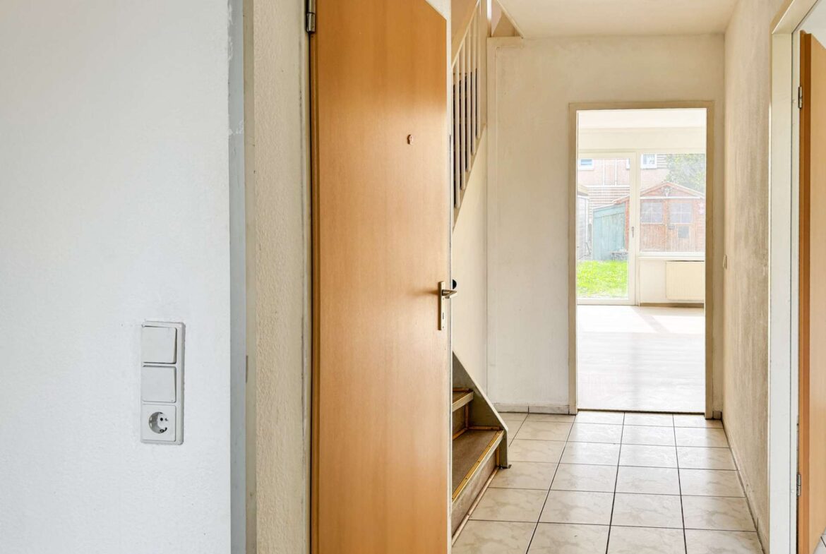 Indoor hallway with white walls, tiled floor, and an open wooden door; stairs visible on the left and a bright room at the end revealing a yard outside.