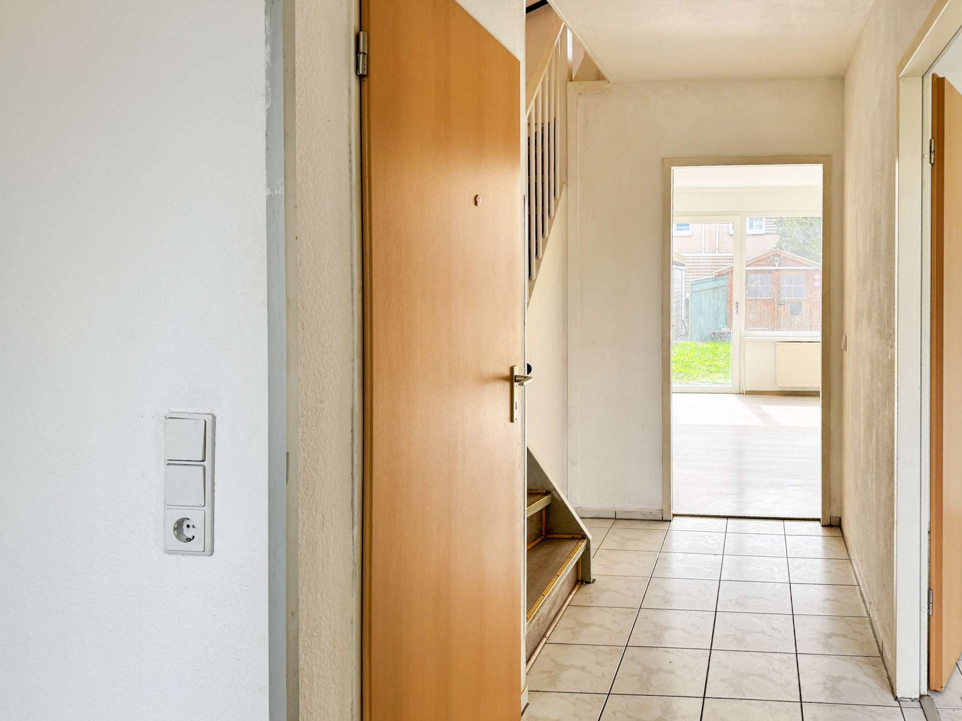 Indoor hallway with white walls, tiled floor, and an open wooden door; stairs visible on the left and a bright room at the end revealing a yard outside.