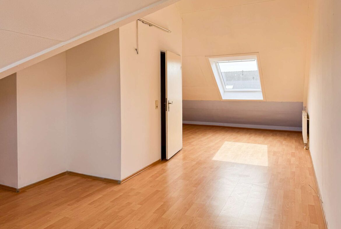 Sunlit empty attic room with a slanted ceiling, skylight window, and light wood floor.