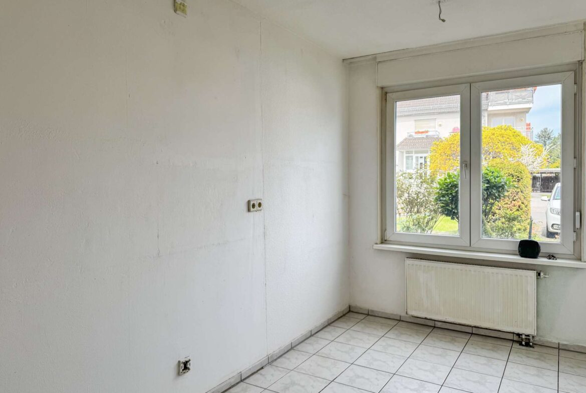 Empty white-walled room with tiled floor and a large window overlooking a garden and street, radiator beneath the window and a few outlets visible on the wall.