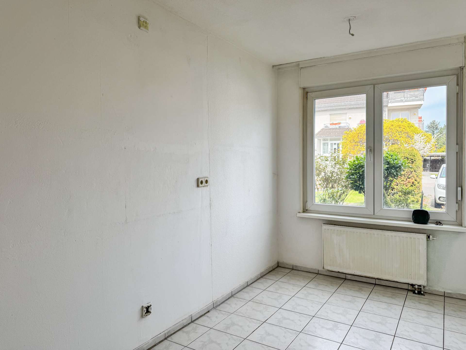 Empty white-walled room with tiled floor and a large window overlooking a garden and street, radiator beneath the window and a few outlets visible on the wall.
