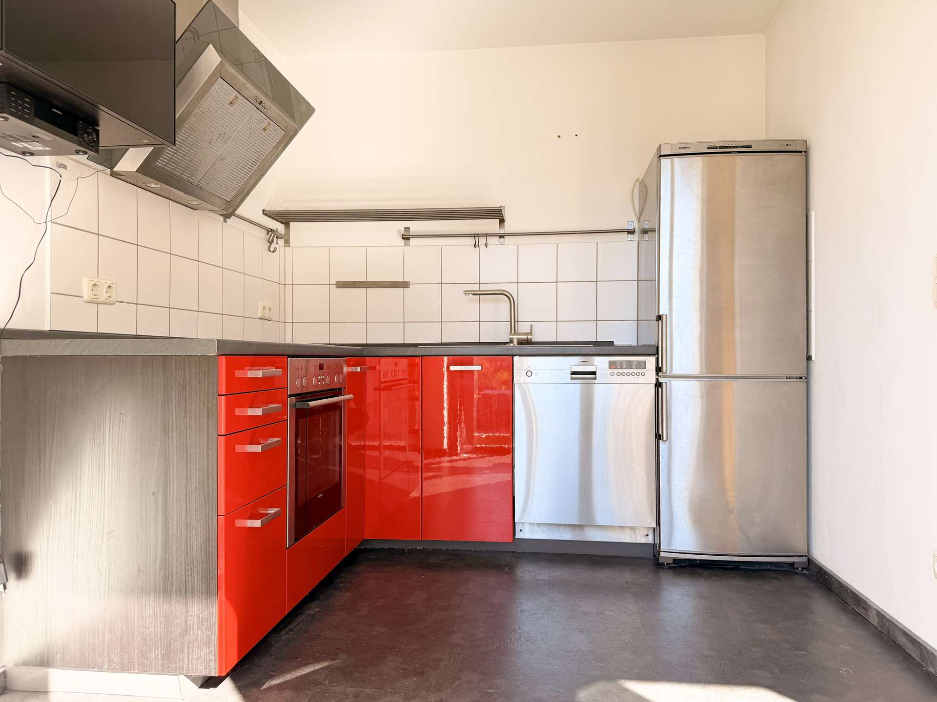 Compact kitchen with glossy red cabinets, stainless oven, and a tall silver refrigerator against a white-tiled backsplash.