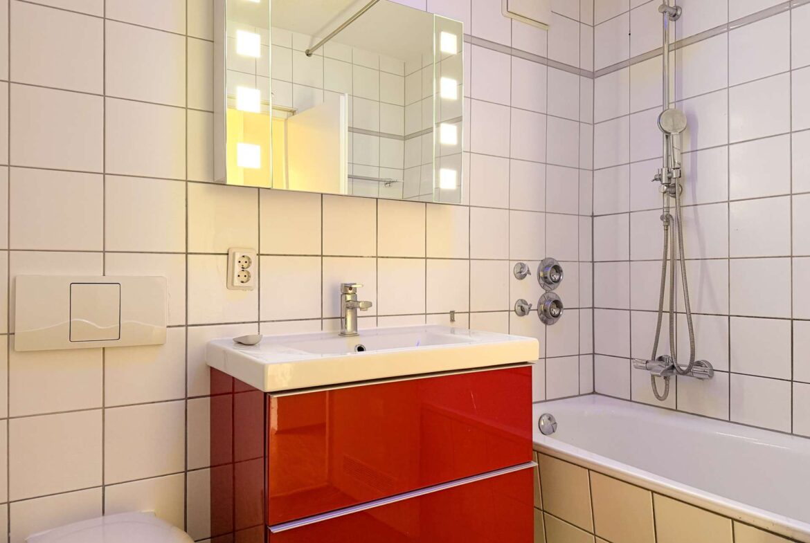 Modern bathroom with white tiled walls, a red glossy vanity under a white sink, and a backlit mirror above the sink.