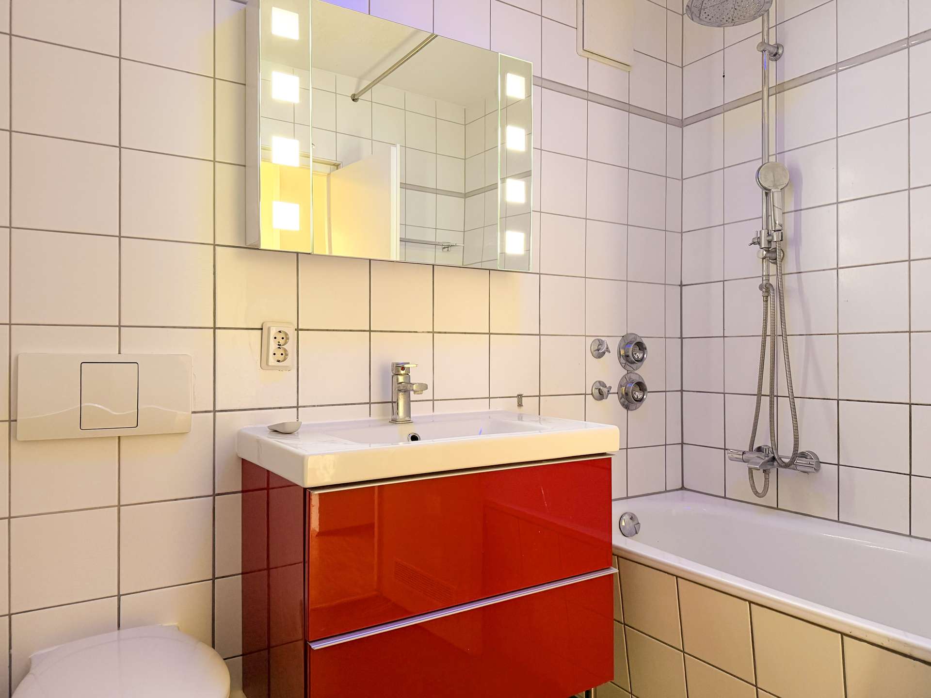Modern bathroom with white tiled walls, a red glossy vanity under a white sink, and a backlit mirror above the sink.