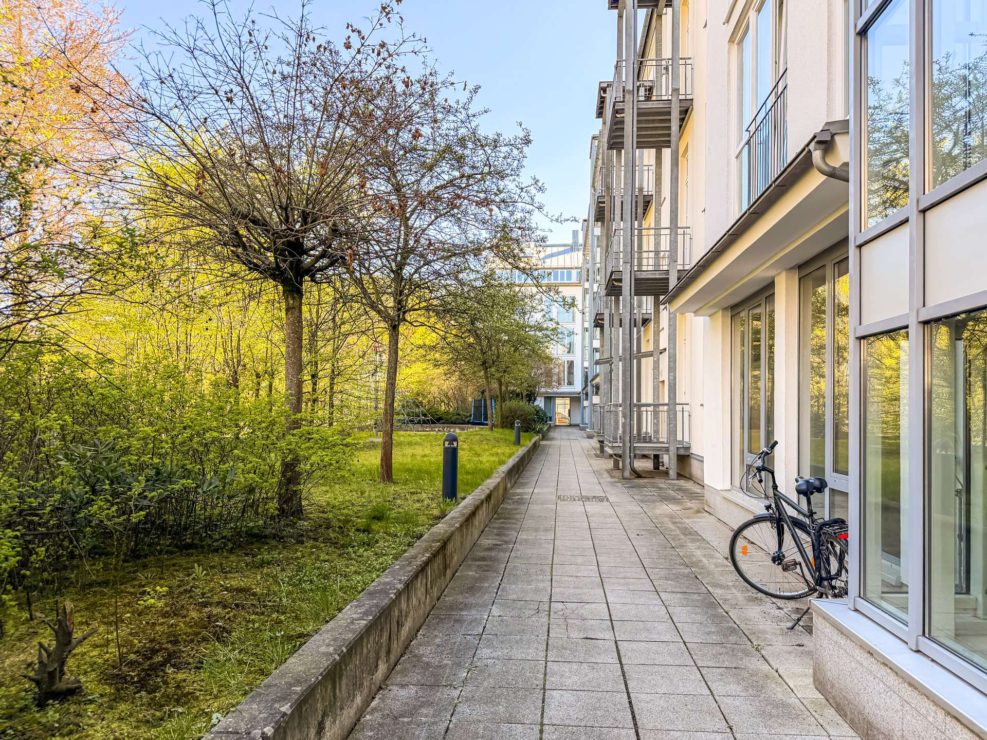 Footpath beside a modern apartment building with glass windows and balconies, bordered by trees and greenery; a bicycle parked by the wall.