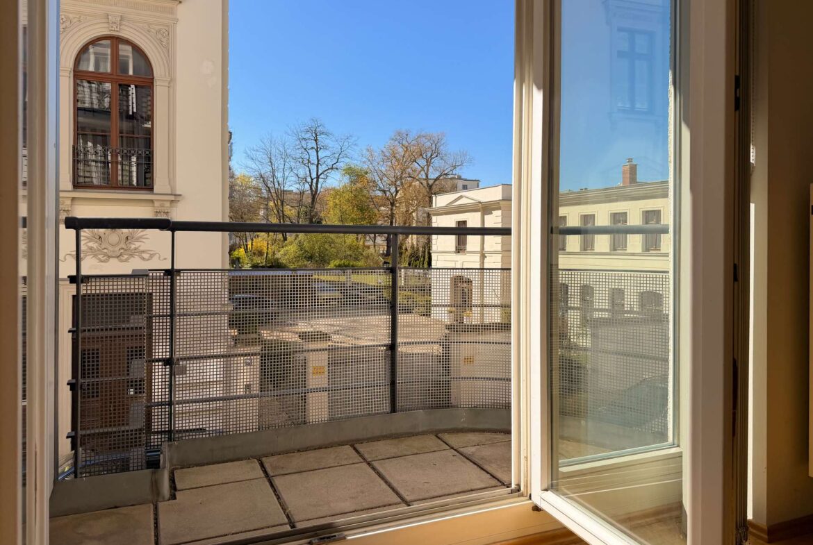 Open balcony door to a small terrace with a metal railing; view of beige buildings, leafless trees, and a bright blue sky.