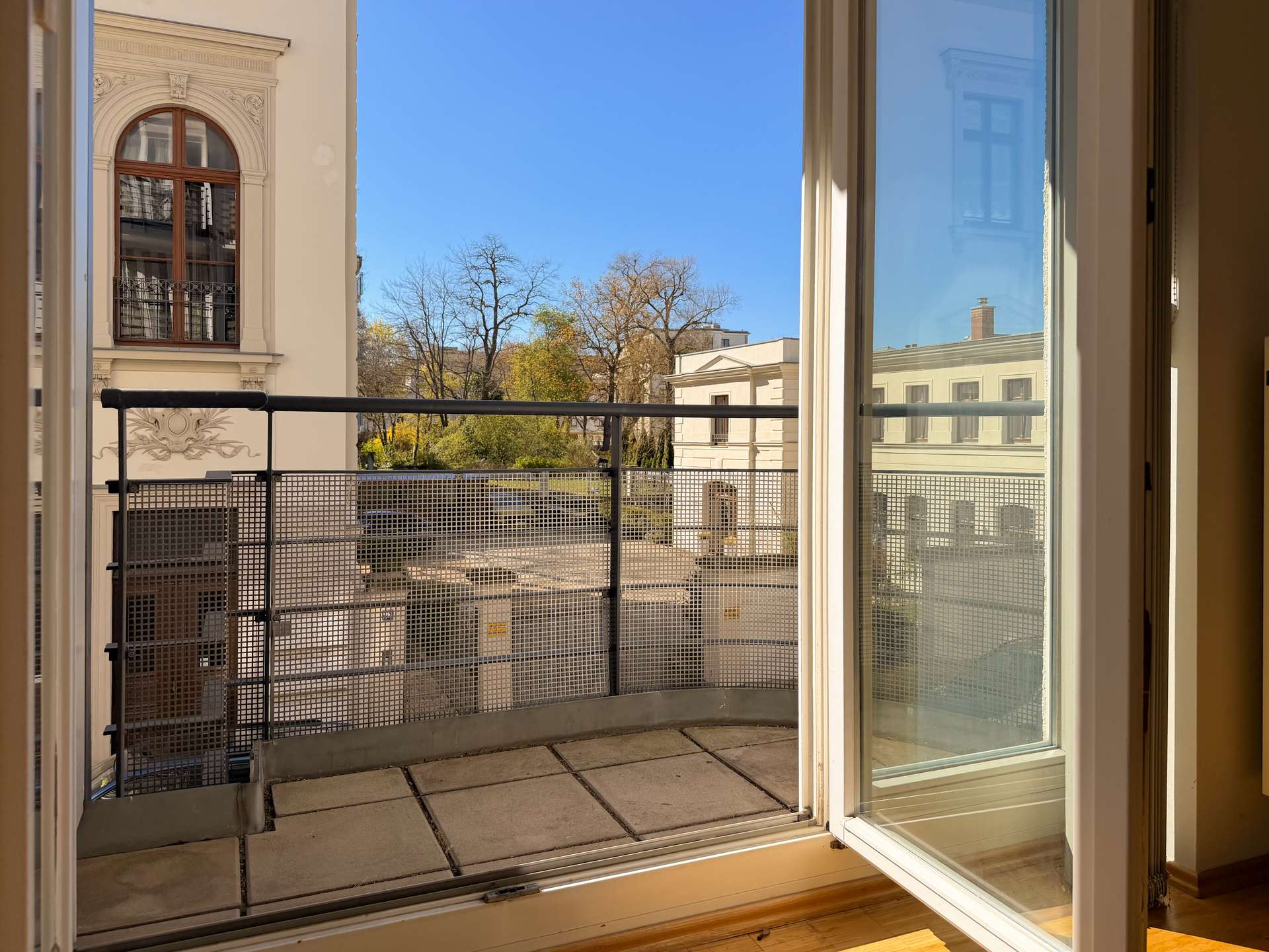 Open balcony door to a small terrace with a metal railing; view of beige buildings, leafless trees, and a bright blue sky.