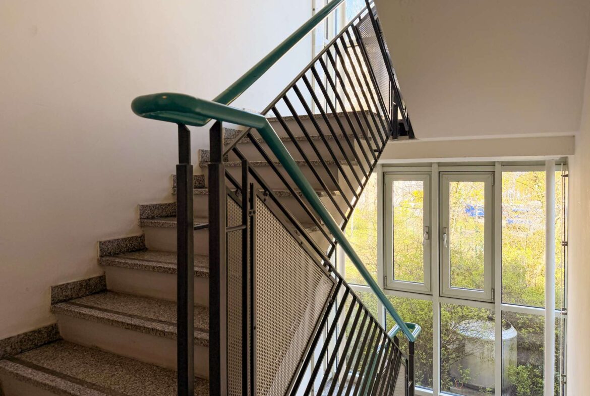 Indoor stairwell with a teal-green handrail and black metal railing; large window lets in daylight and shows trees outside.