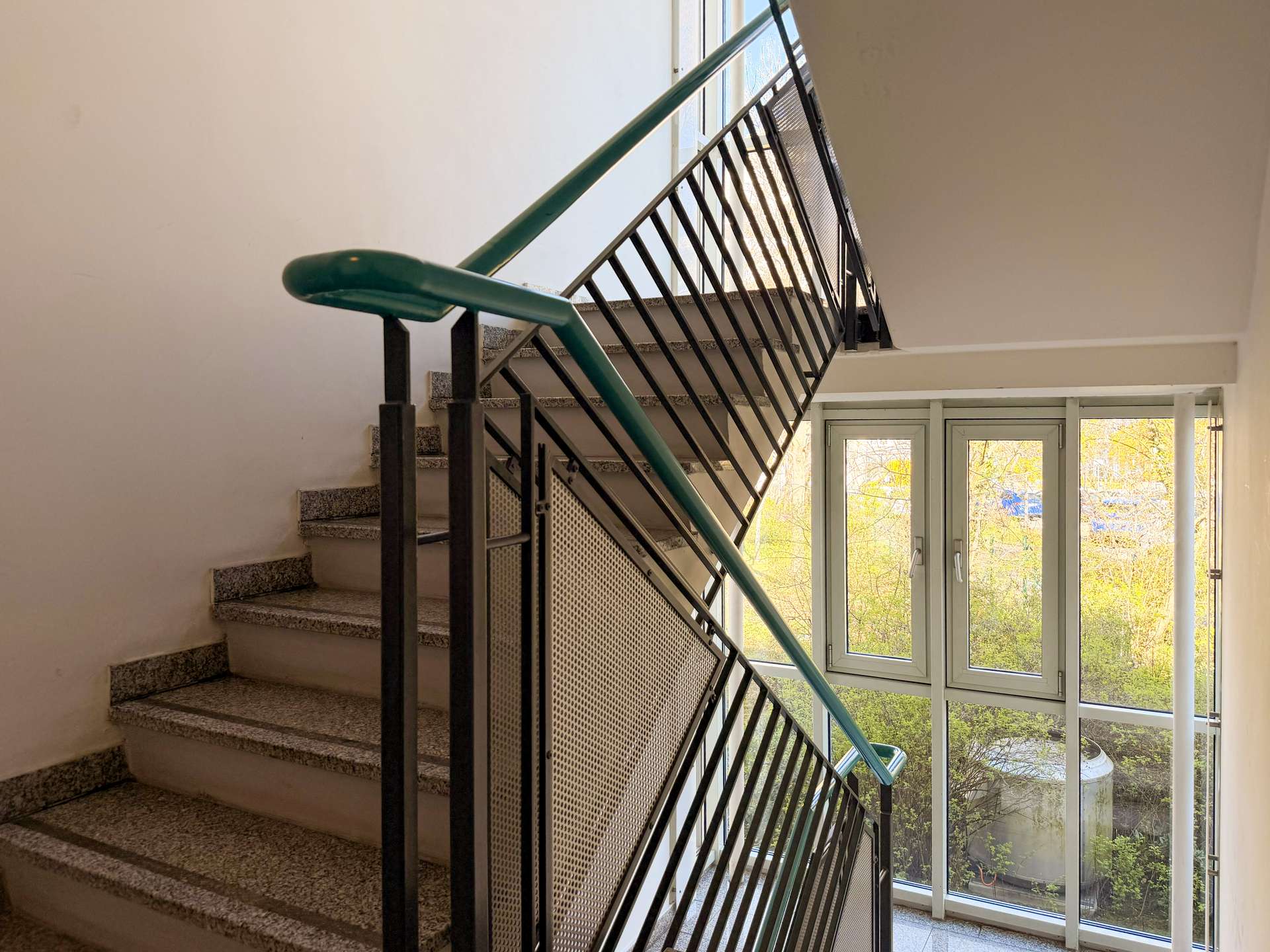 Indoor stairwell with a teal-green handrail and black metal railing; large window lets in daylight and shows trees outside.