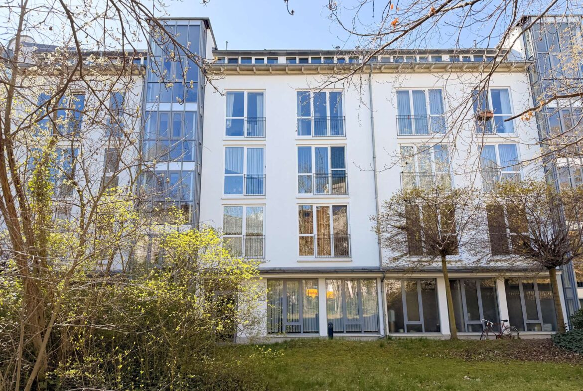 White multi-story residential building with large glass balconies, seen through leafless trees in a sunny courtyard.