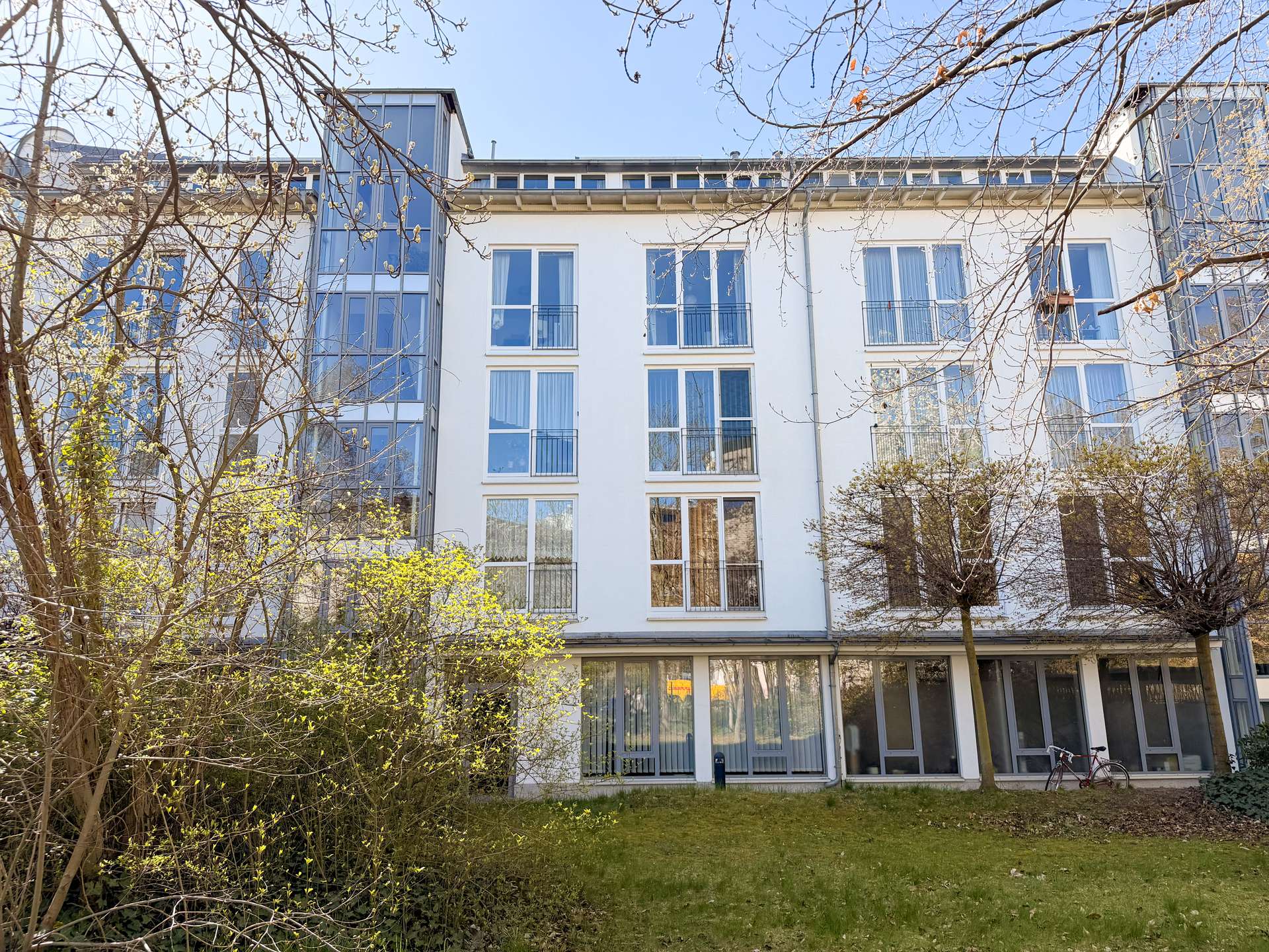 White multi-story residential building with large glass balconies, seen through leafless trees in a sunny courtyard.