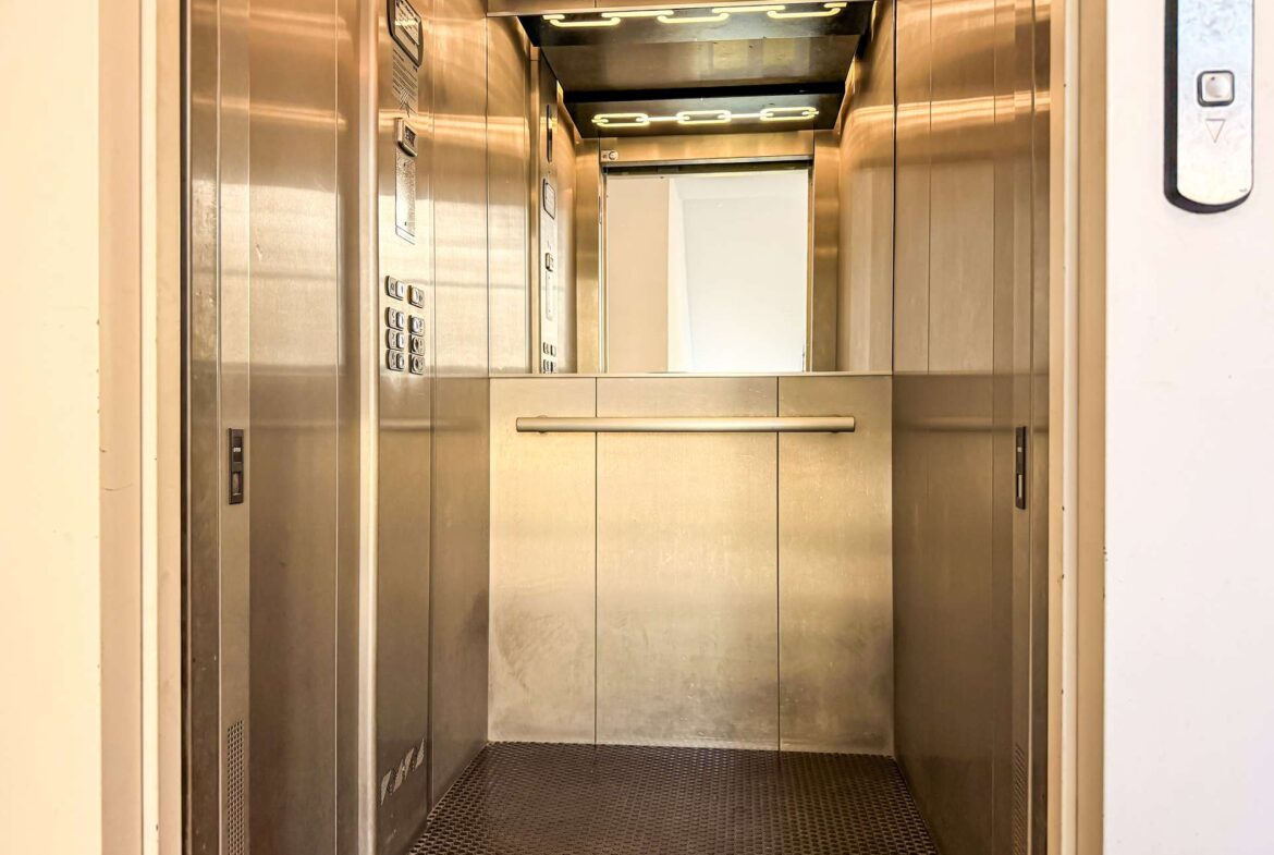 Empty stainless-steel elevator cab with left control panel, overhead lights, and a mid-height horizontal handrail.