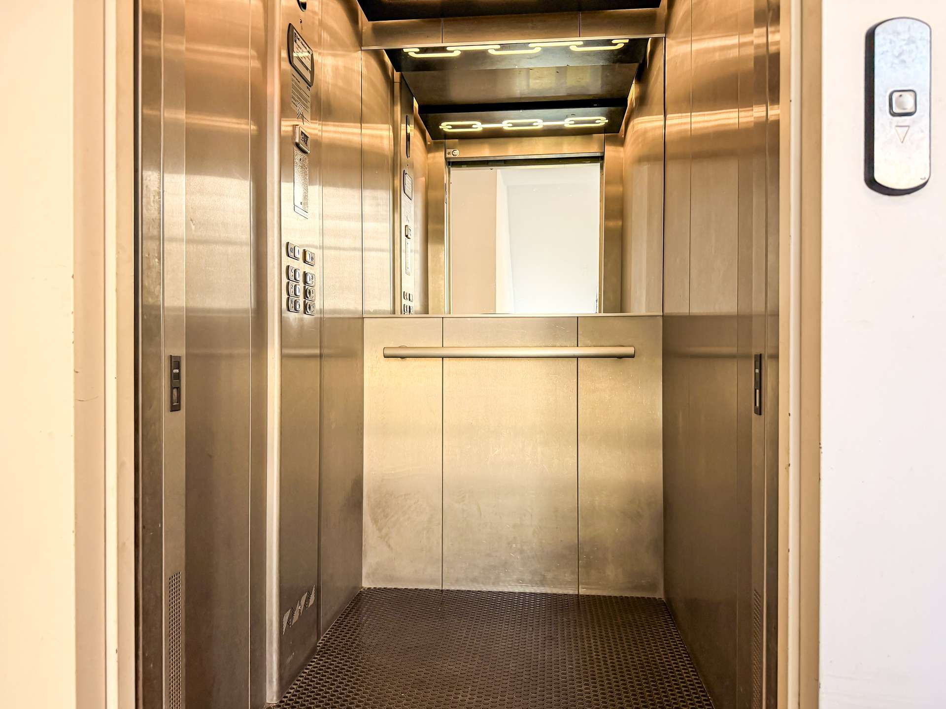 Empty stainless-steel elevator cab with left control panel, overhead lights, and a mid-height horizontal handrail.