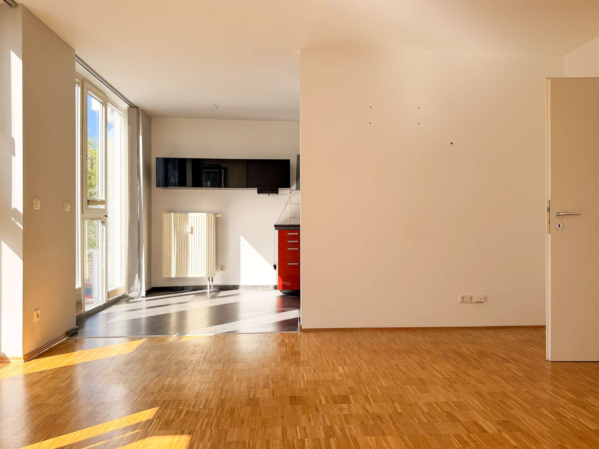 Sunlit interior with large left-side windows, a radiator, and a red tool cabinet against the back wall in an empty room.