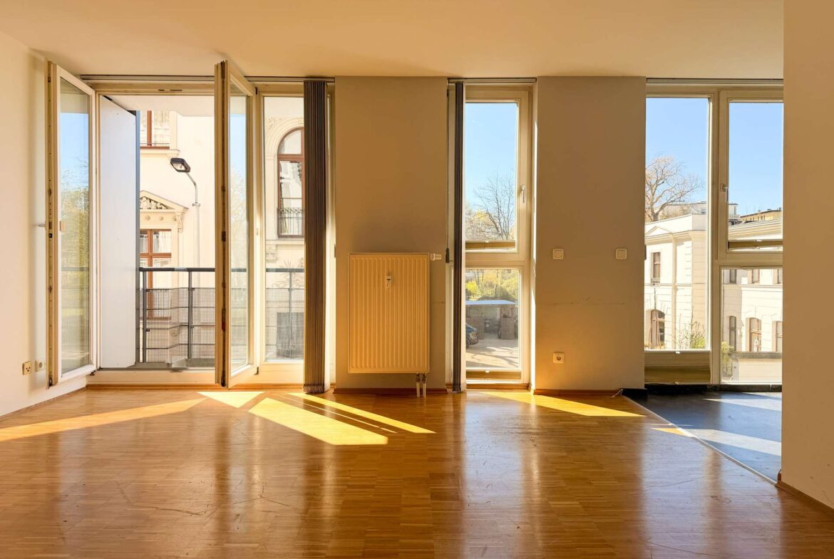 Sunlit empty living room with tall windows and an open balcony door, casting warm light on a wooden floor.