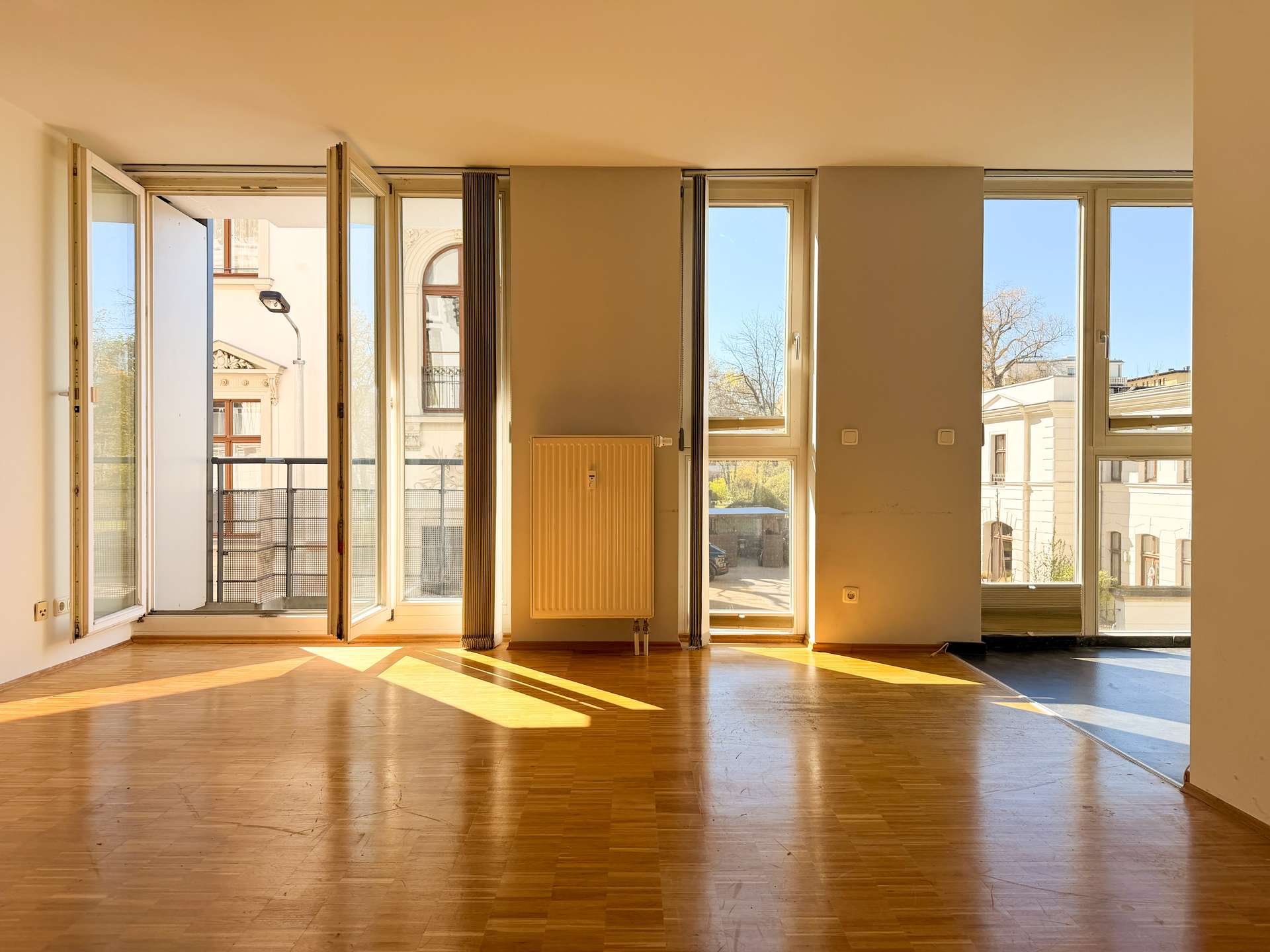 Sunlit empty living room with tall windows and an open balcony door, casting warm light on a wooden floor.