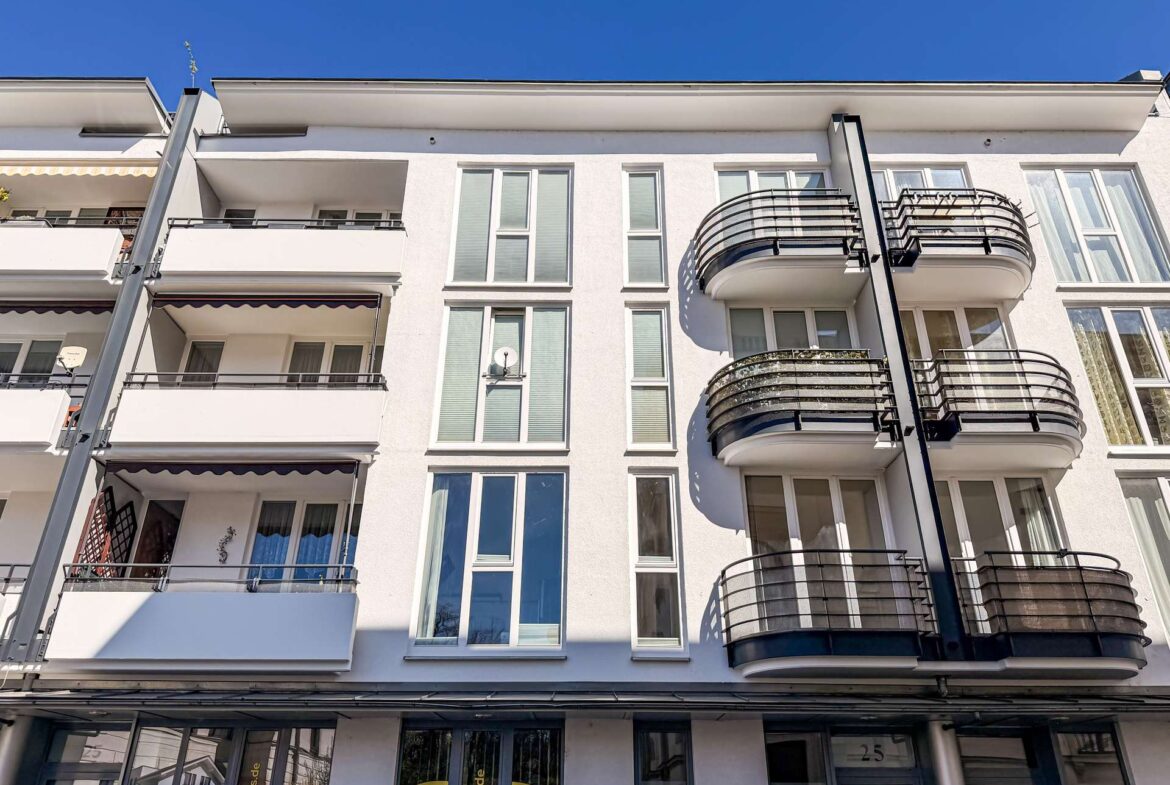White modern apartment building with tall windows and curved black balconies on the right, under a clear blue sky.