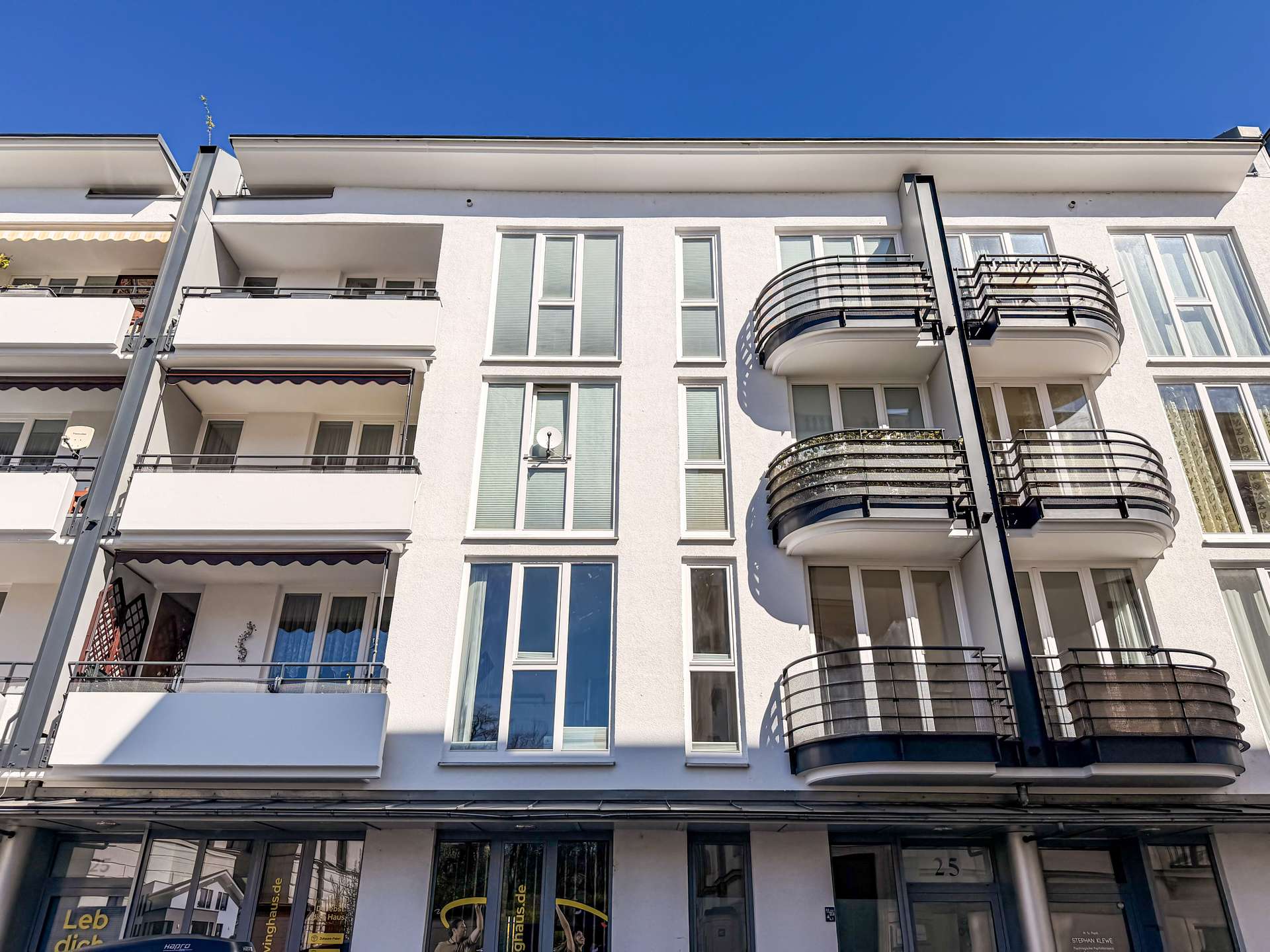 White modern apartment building with tall windows and curved black balconies on the right, under a clear blue sky.
