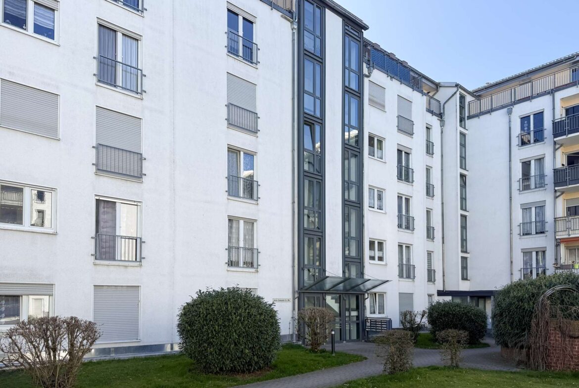 White residential building with multiple balconies and glass entrance, seen from a green courtyard.