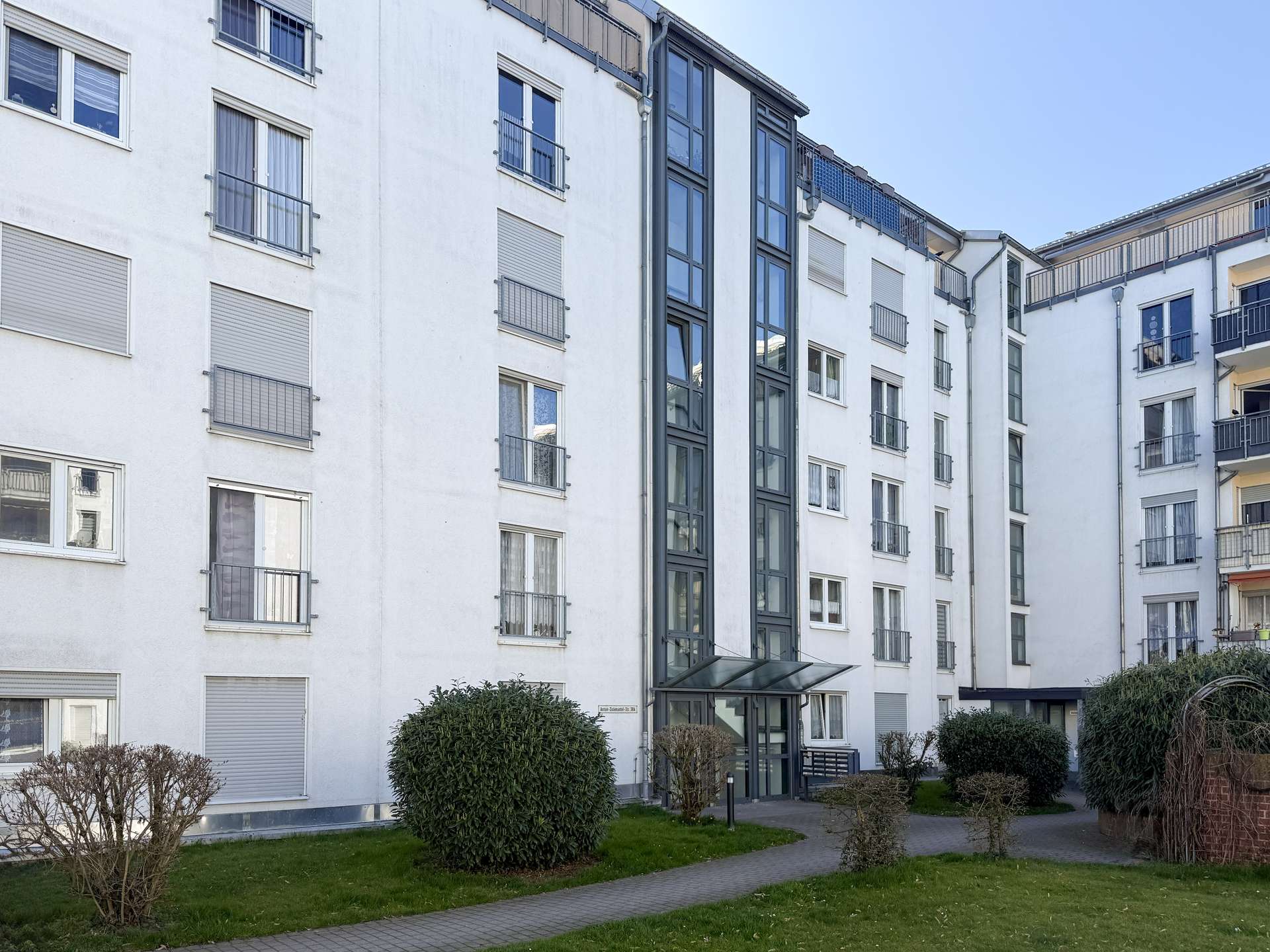 White residential building with multiple balconies and glass entrance, seen from a green courtyard.
