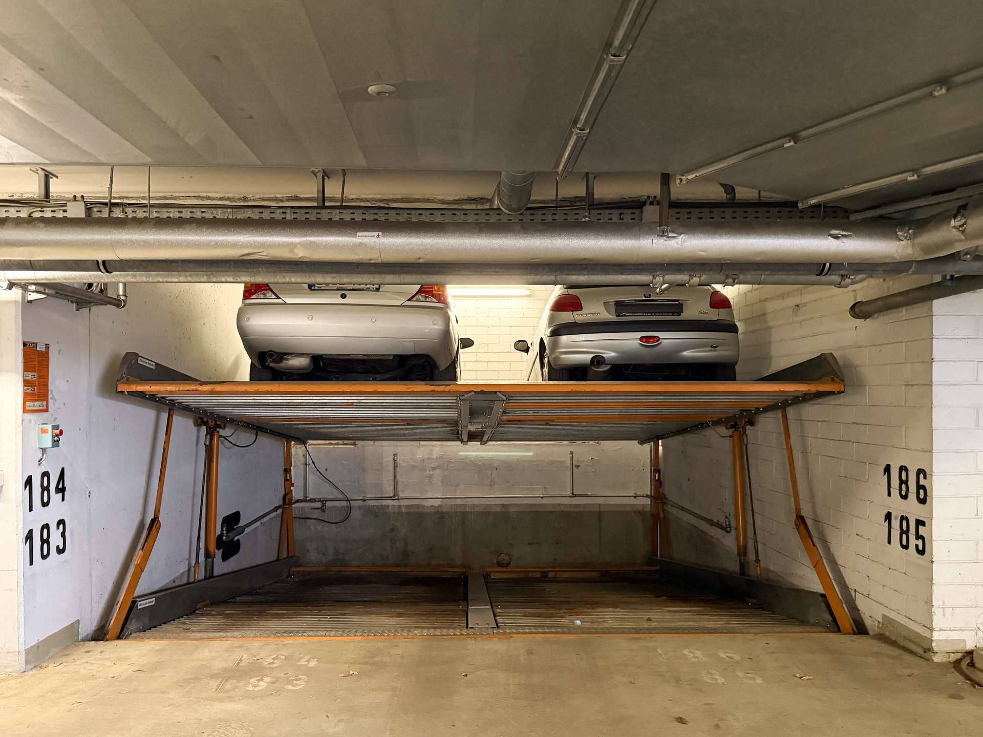 Underground parking garage with two silver cars on a raised vehicle lift between white brick walls and exposed pipes.