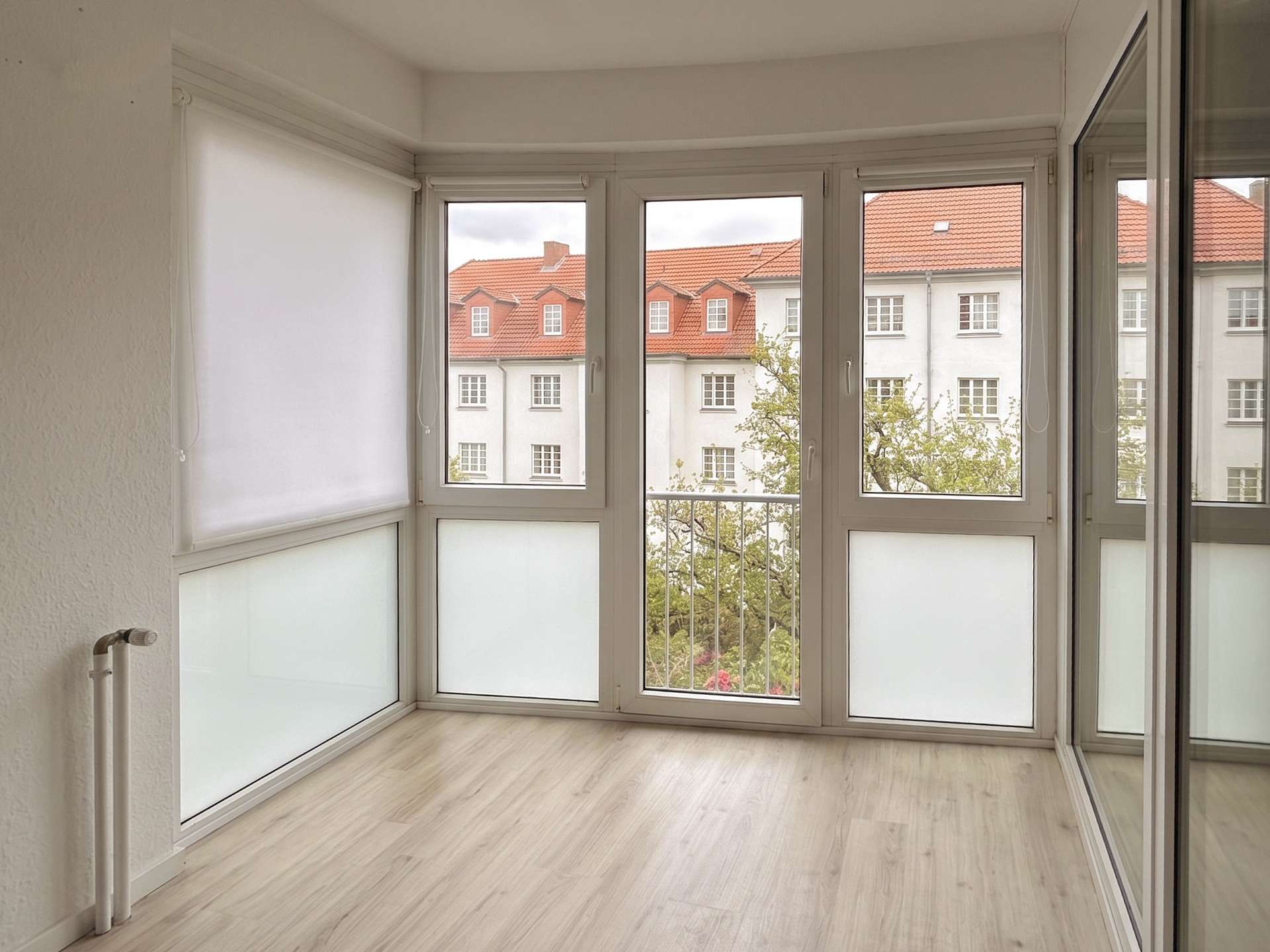 Bright enclosed balcony with white window frames; view of red-tiled rooftops and trees outside.