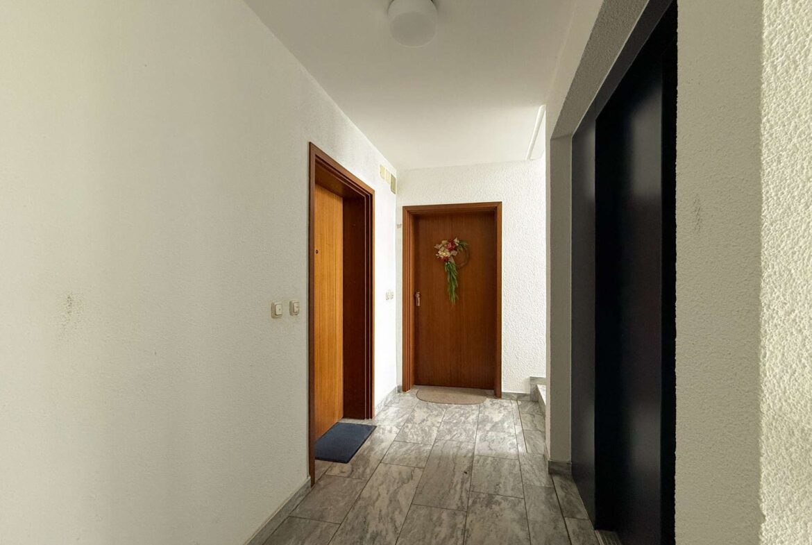 Hallway in a residential building with several wooden doors and a wreath on the far door, tiled gray floor, white walls.