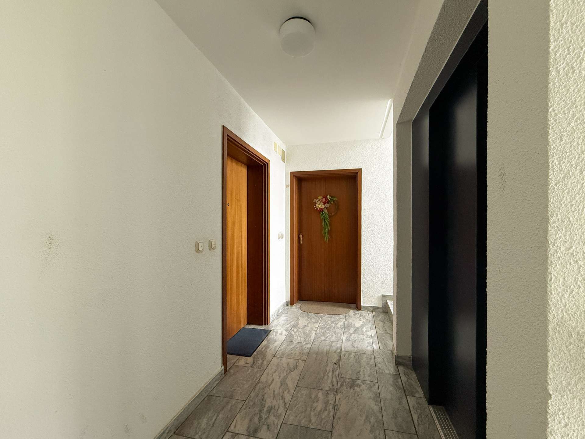 Hallway in a residential building with several wooden doors and a wreath on the far door, tiled gray floor, white walls.