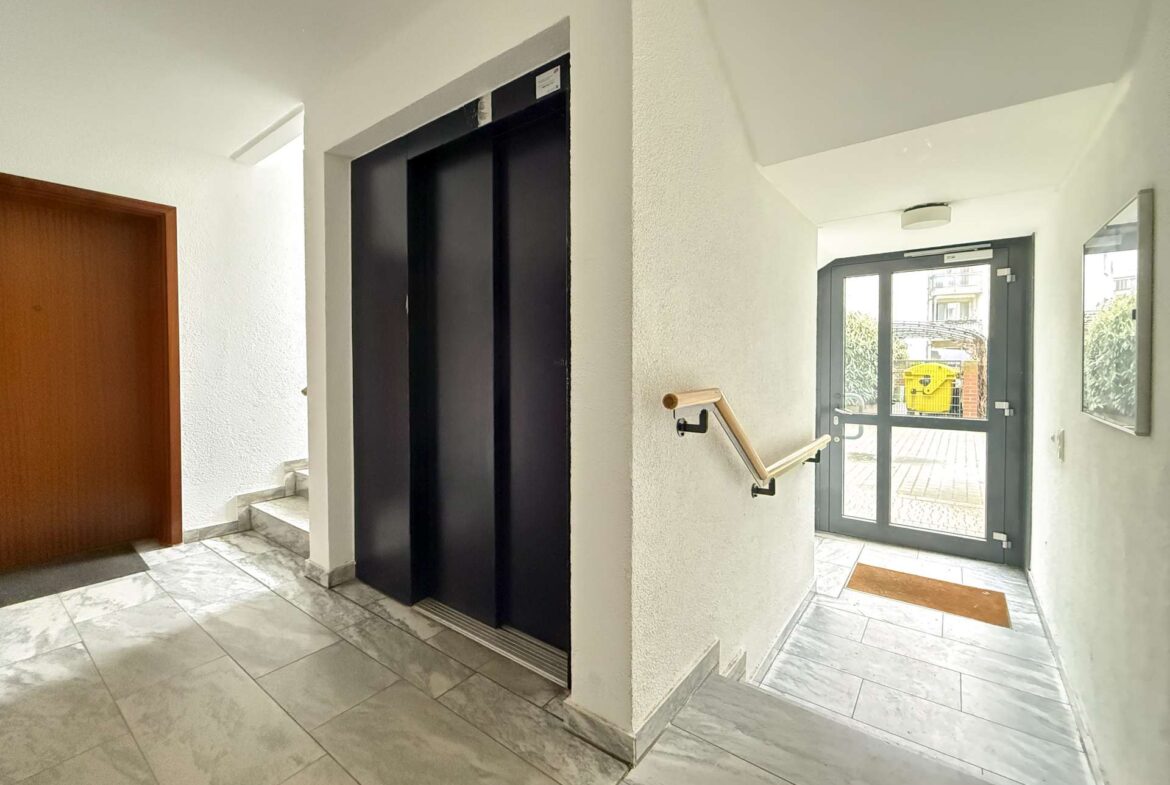 Lobby hallway with a closed black elevator and a staircase nearby, marble tile floor, and a glass exit door at the end of the hall.